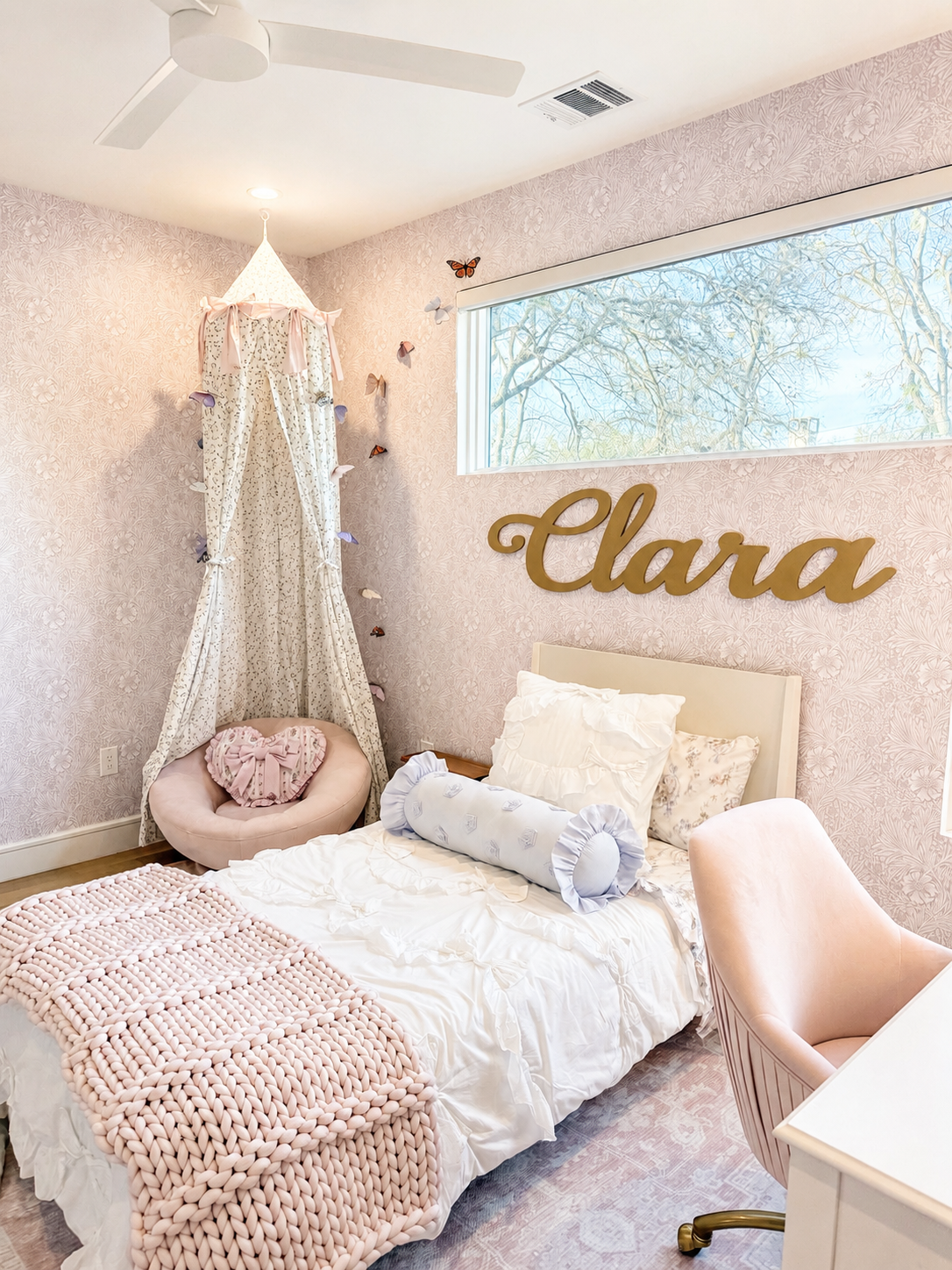 A cozy bedroom with pink floral wallpaper, a white bed with decorative pillows and a chunky pink blanket, a pink vanity chair, a pink canopy with butterfly decorations, a large window with a tree outside, and the name 'Clara' in gold lettering on the wall.
