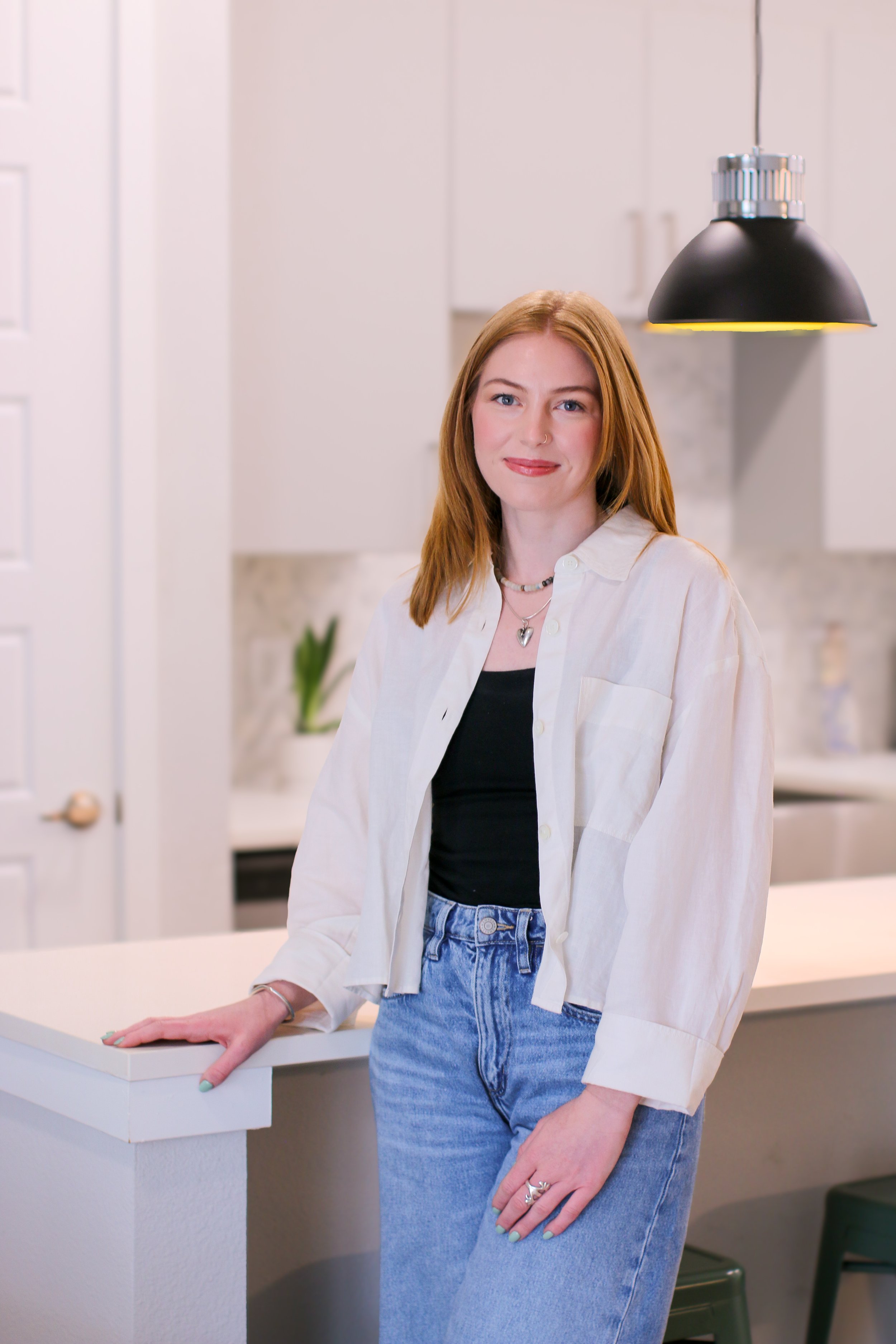 A young woman with red hair, wearing a white button-up shirt over a black top and jeans, standing in a bright, modern kitchen.