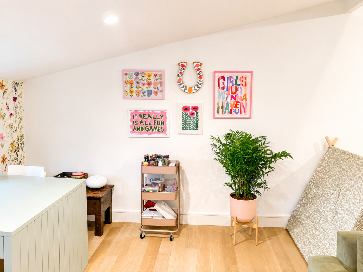 Decorative wall with pink frames featuring colorful artwork and a horseshoe, a potted plant on a small stand, a mobile storage cart with art supplies, a bench, and a piece of patterned art panel leaning against the wall.