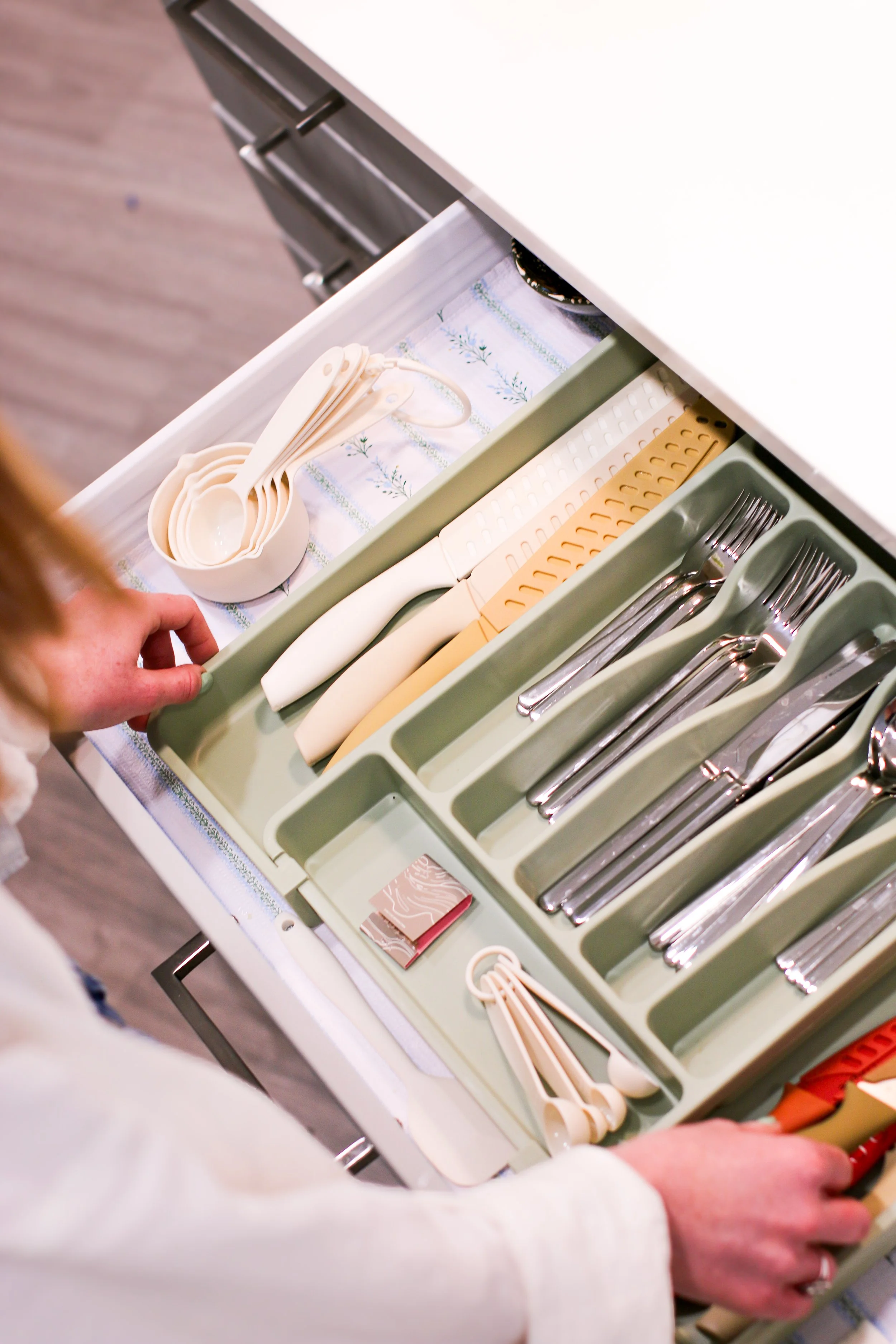 Open kitchen drawer containing white measuring spoons, some beige utensils, small pink and beige paper notepad, and multiple compartments with silver forks and knives.
