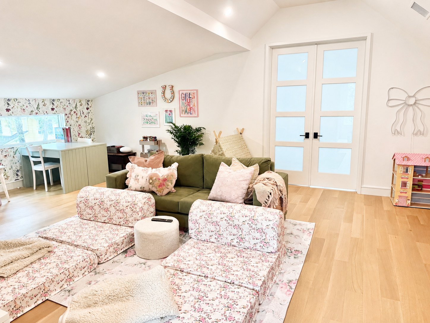 Living room with a green sofa, floral pillows, pink velvet chairs, and various decorative items inside a bright space with wooden flooring and a white door.