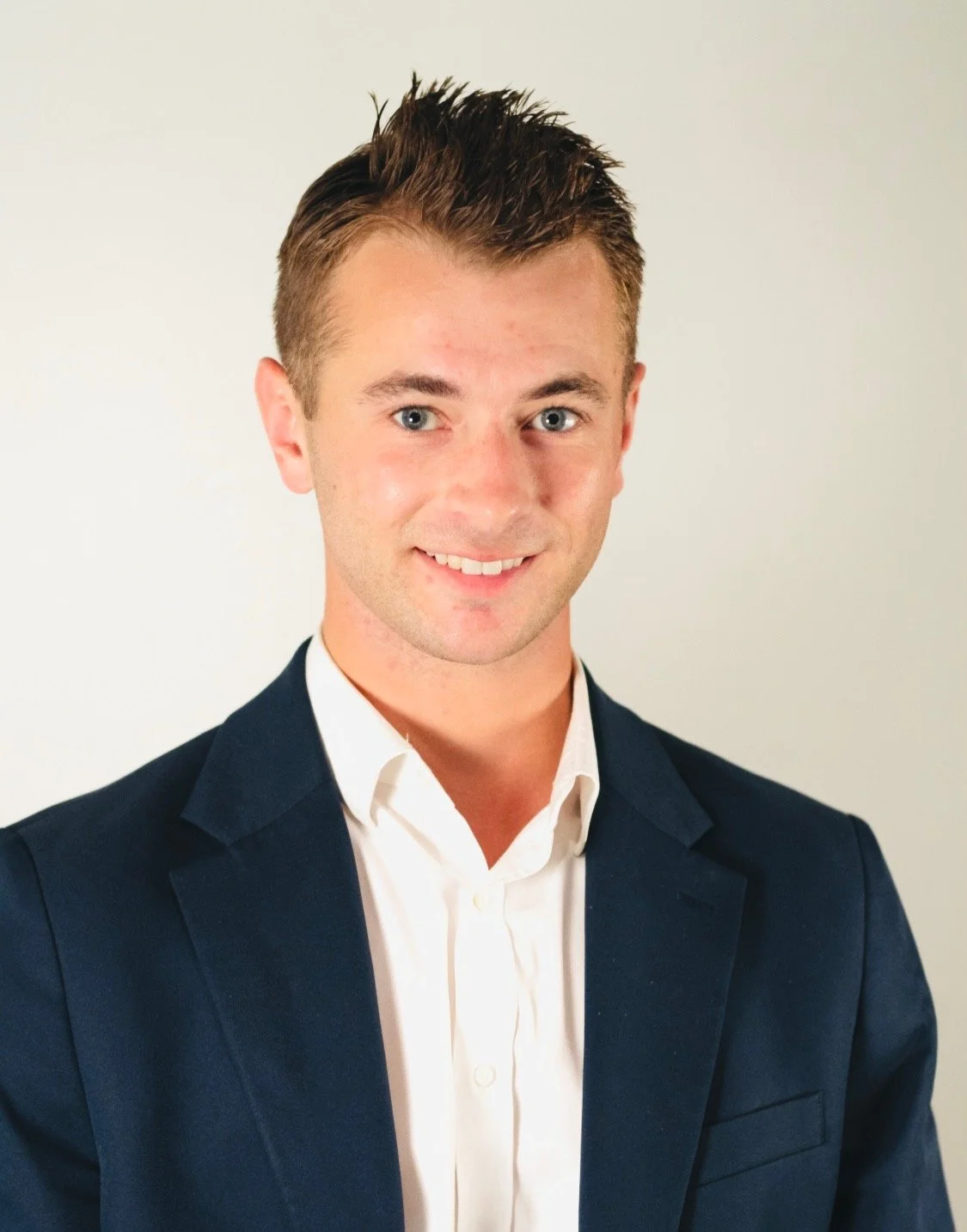 A young man with short, styled brown hair and blue eyes, wearing a dark blue suit jacket and a white dress shirt, smiling against a plain light-colored background.