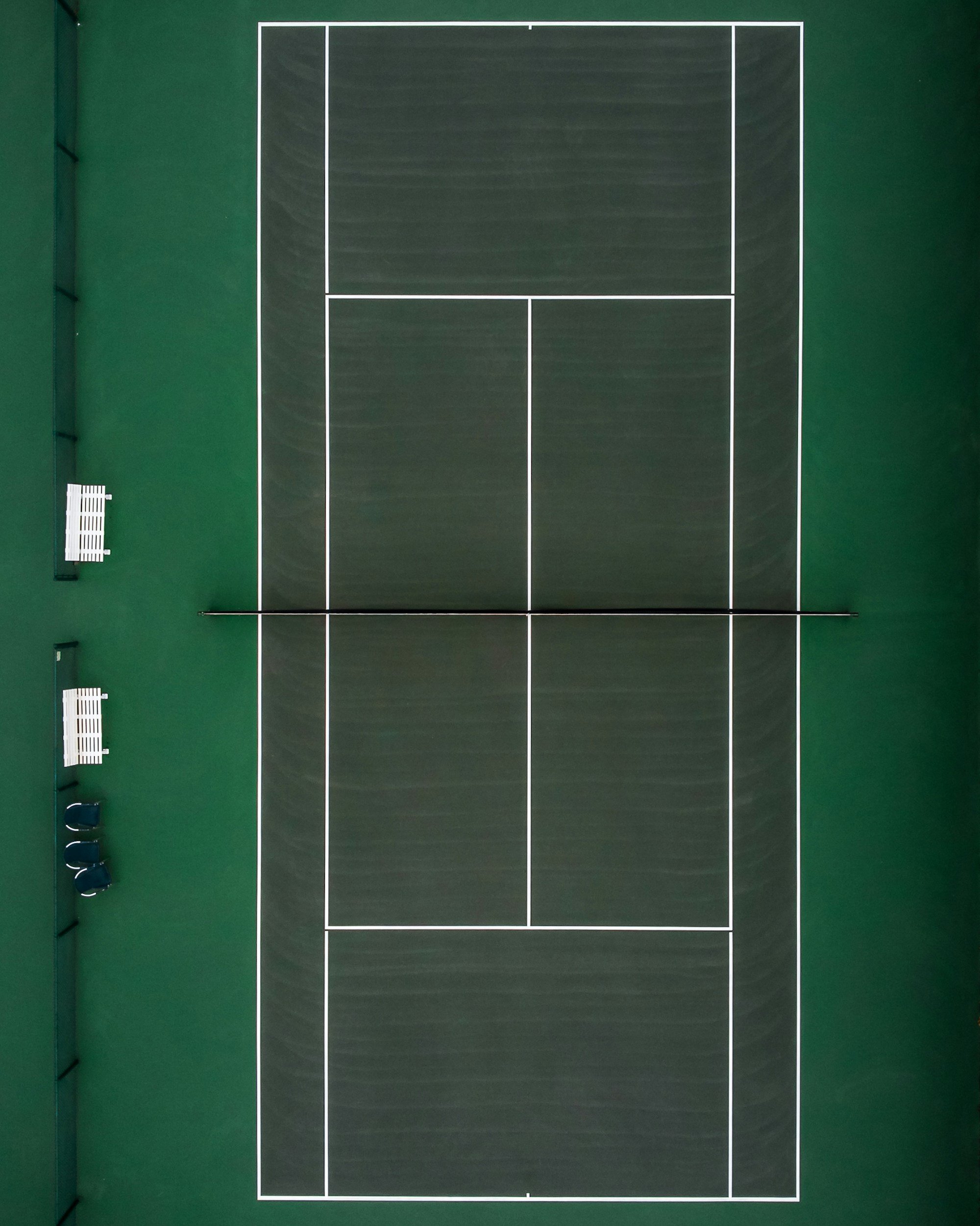 A tennis court with green surface, white lines, net in the middle, and empty benches and chairs on the sides.