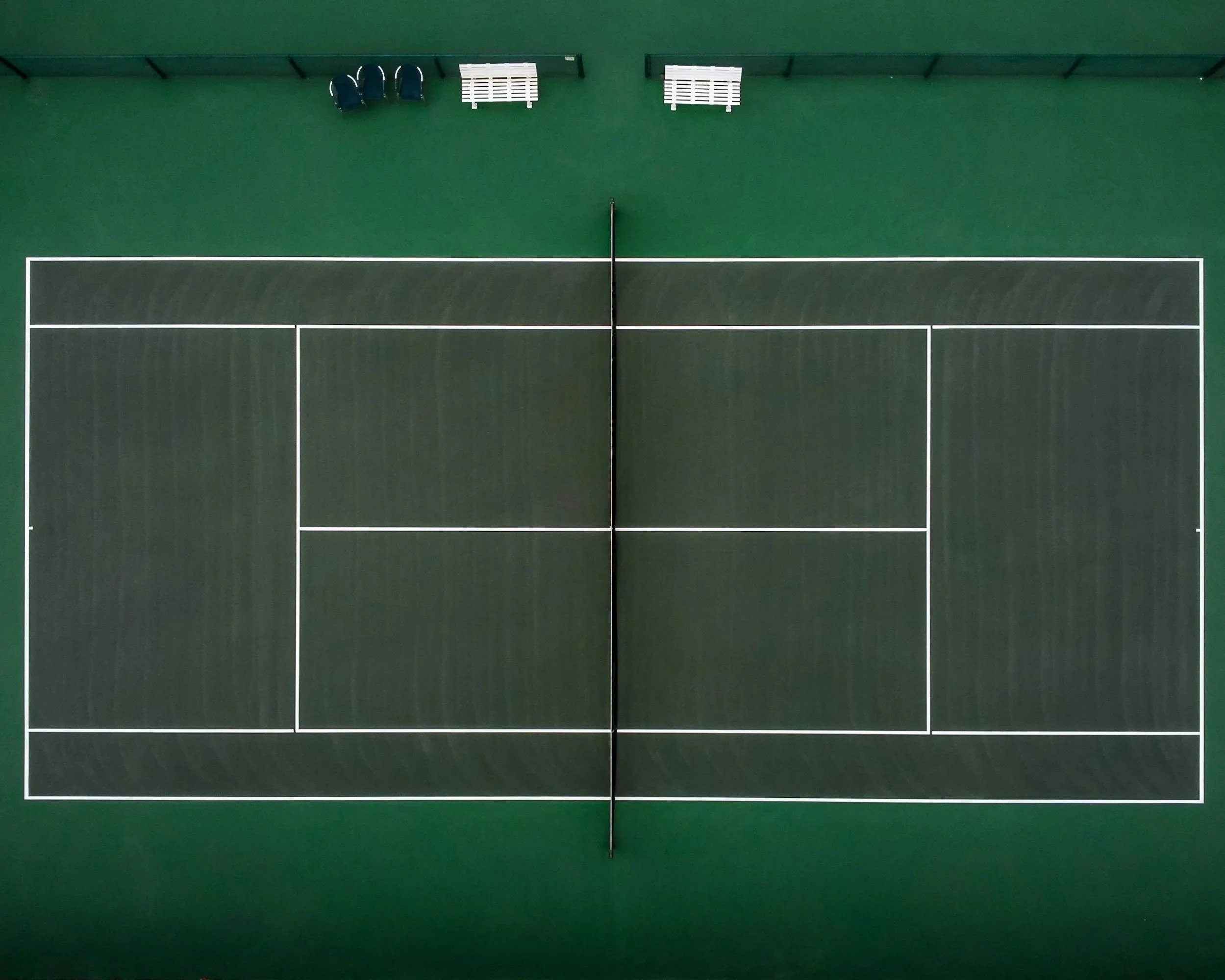 Overhead view of a green tennis court with white boundary and service lines and a net in the center, with three chairs and two white benches along the top.