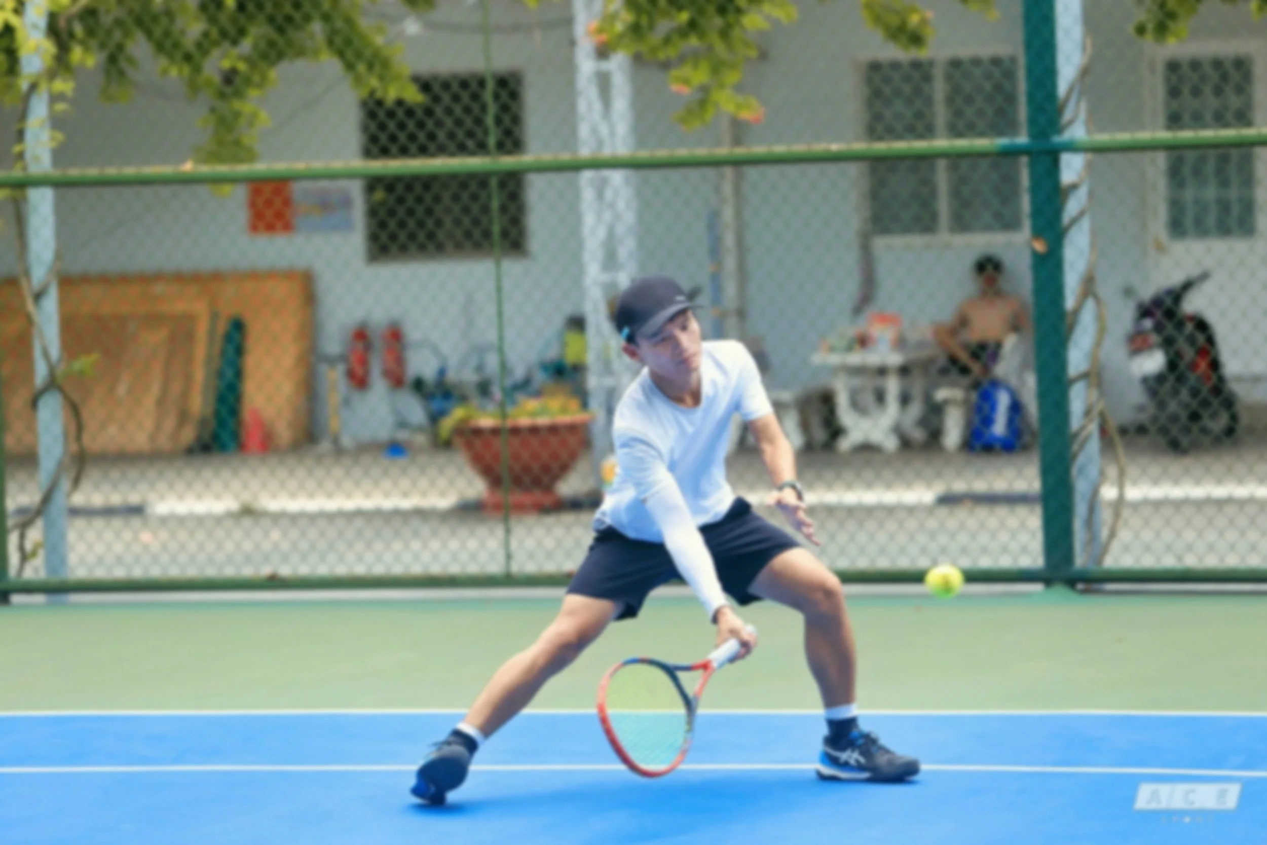 A young man playing tennis on an outdoor court, preparing to hit a tennis ball with a racket. He is wearing a black cap, white long-sleeve shirt, black shorts, and black shoes. The court has a blue playing surface with surrounding green fencing.