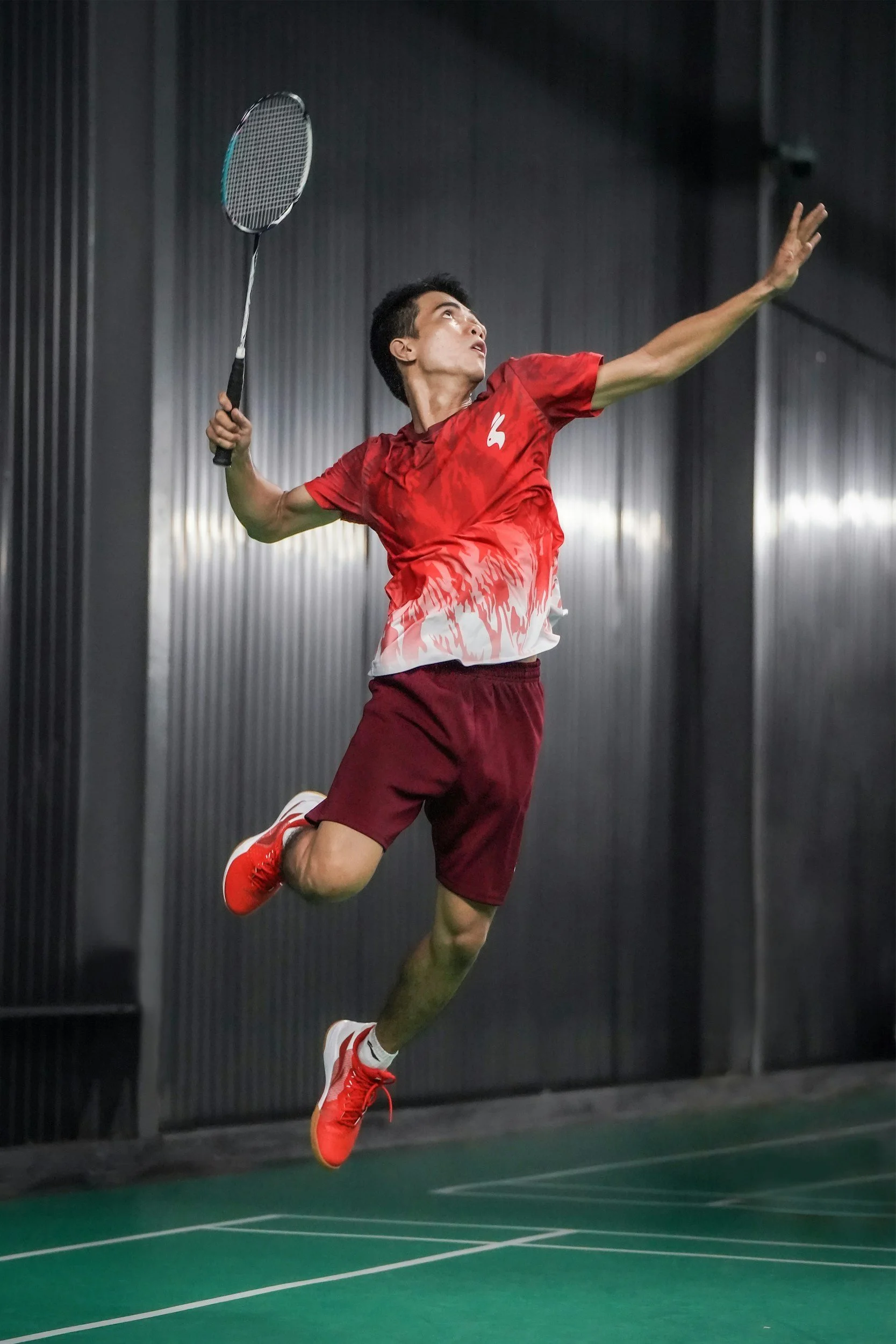 A male badminton player in a red shirt and shorts jumps to hit the shuttlecock with his racket indoors.