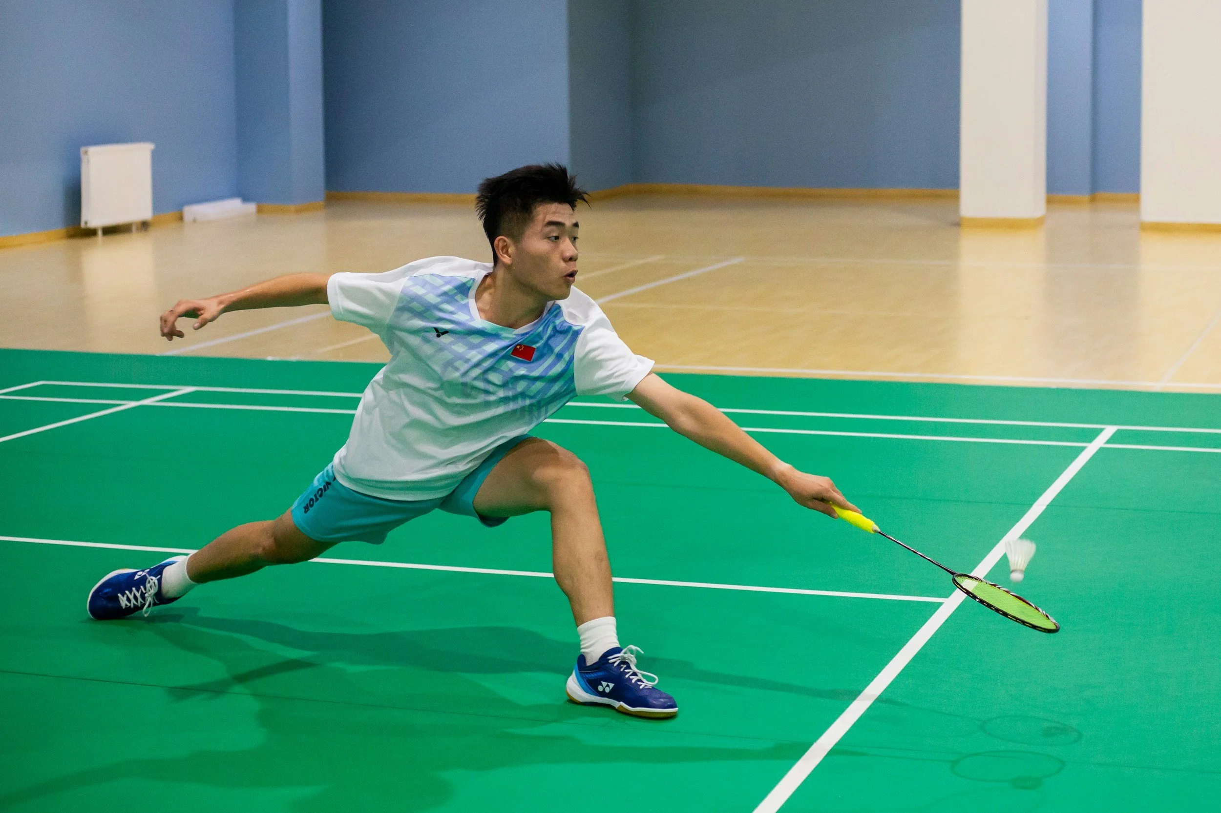 A young man playing badminton indoors, lunging forward with his right hand extended, holding a badminton racket, ready to hit a shuttlecock.