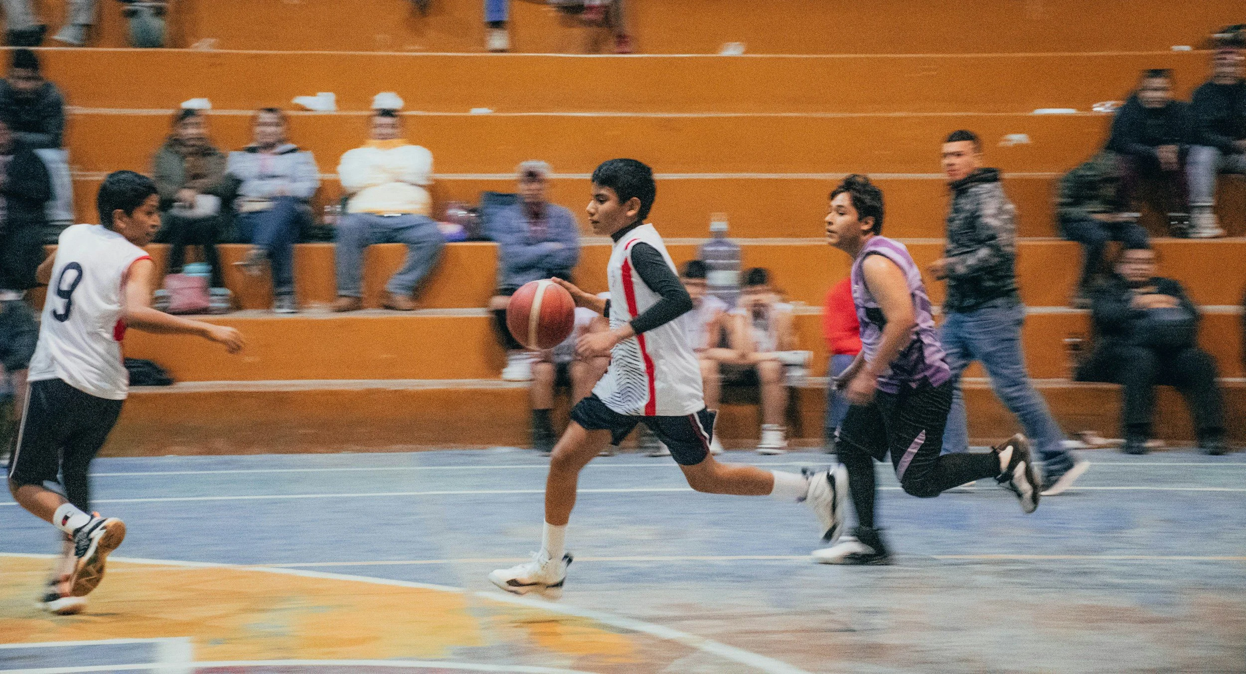 Young boys playing basketball in an indoor gym with spectators seated on bleachers in the background.