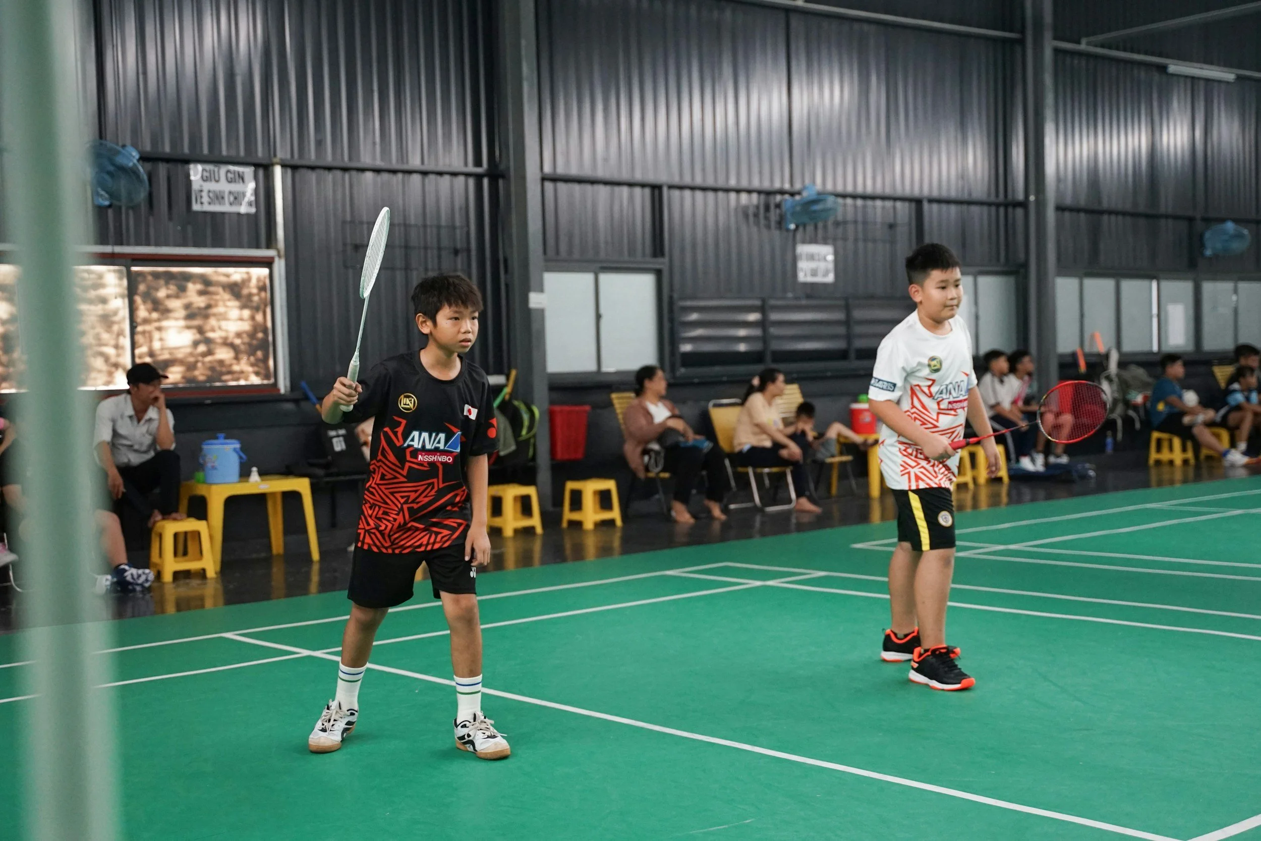 Two young boys playing badminton on an indoor court with spectators seated along the side.