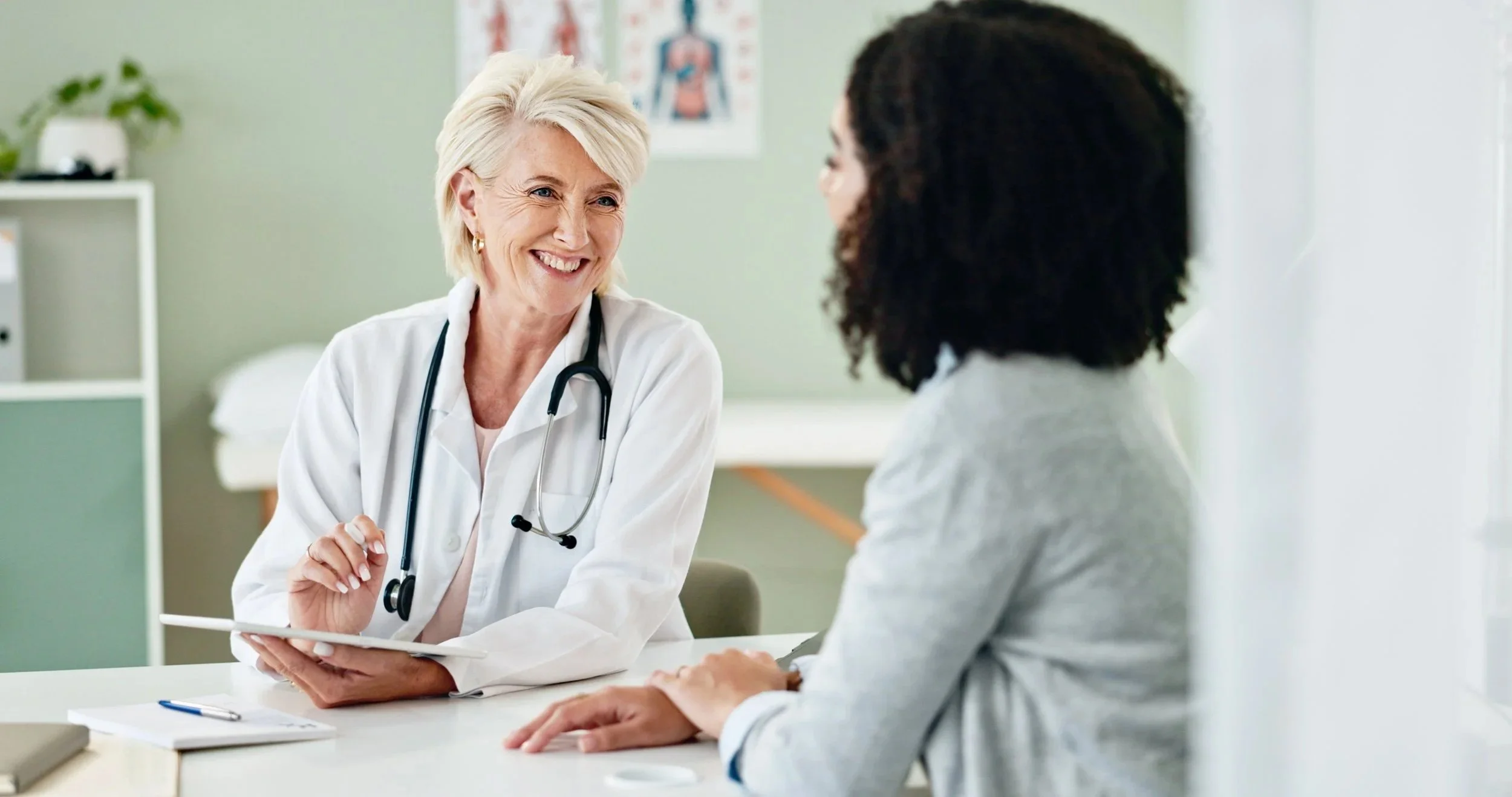 A female doctor with short blonde hair, smiling, wearing a white coat and stethoscope, talking to a patient with dark curly hair in a medical office.