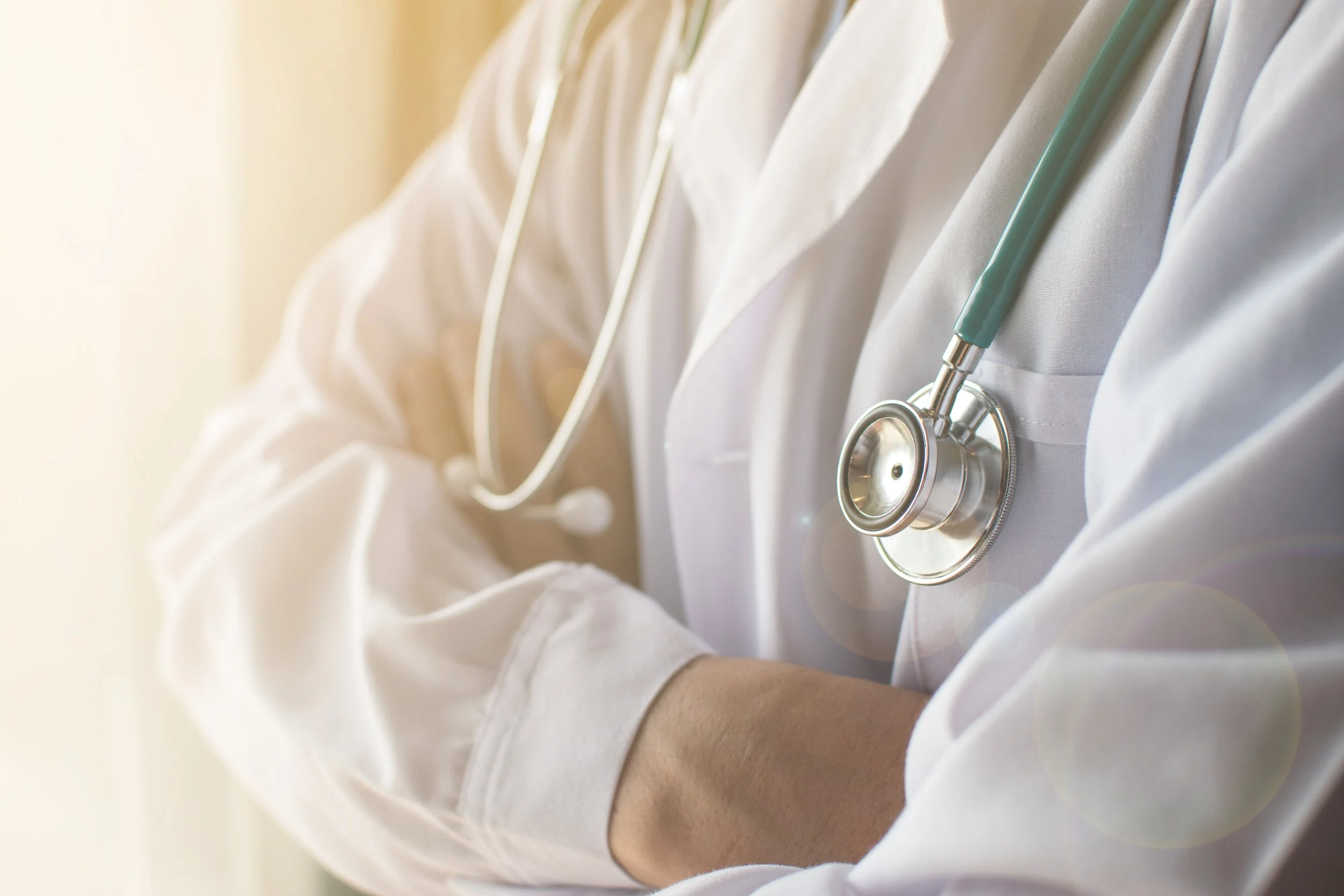 Close-up of a doctor in a white lab coat with a stethoscope around their neck, arms crossed, in a well-lit setting.