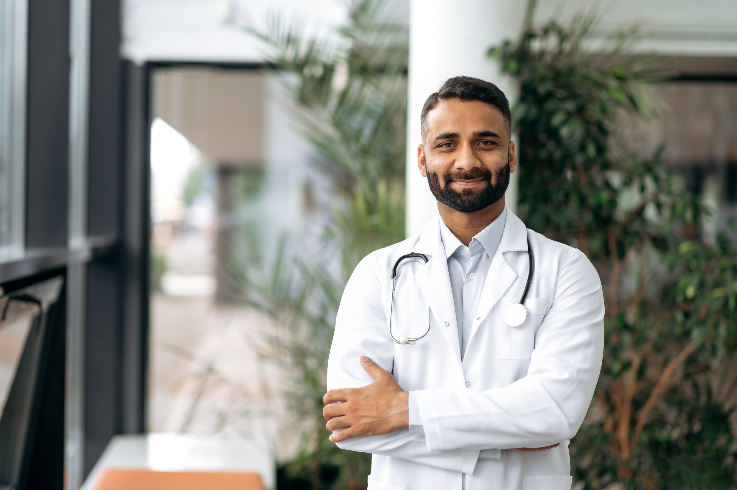 A smiling male doctor in a white coat with a stethoscope around his neck standing in a modern office or hospital corridor with plants in the background.