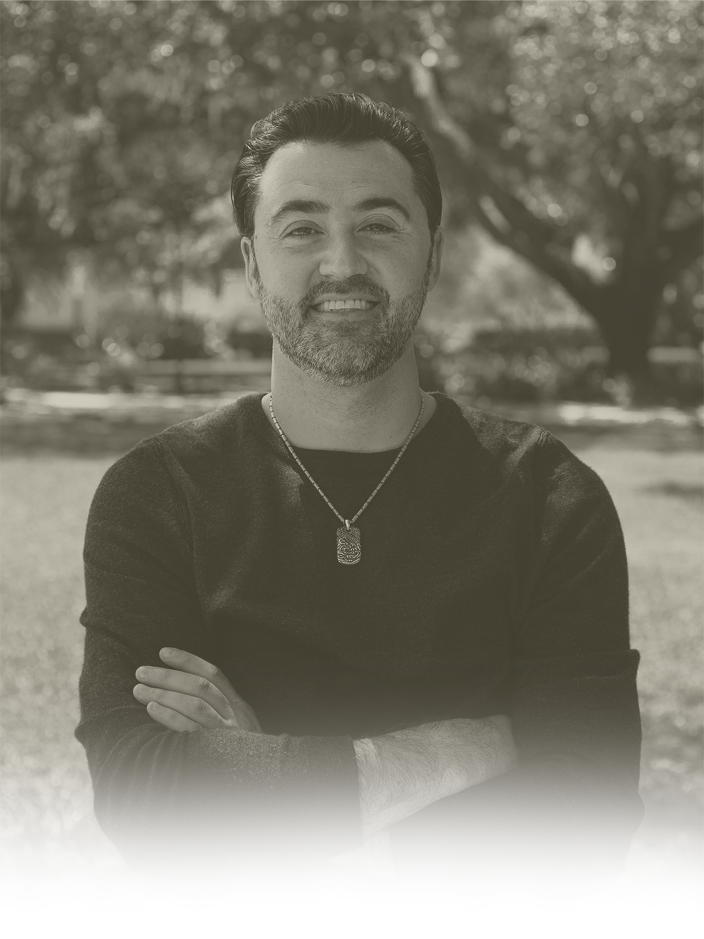 A man with a beard and mustache, smiling with arms crossed, standing outdoors with trees in the background, wearing a black shirt and a necklace.