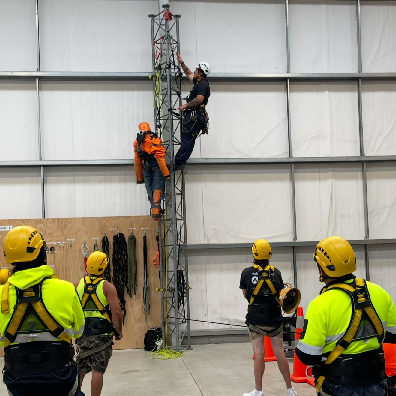 A group of people wearing helmets and safety gear at an indoor training facility watching two individuals practicing climbing a tall metal tower using safety harnesses.