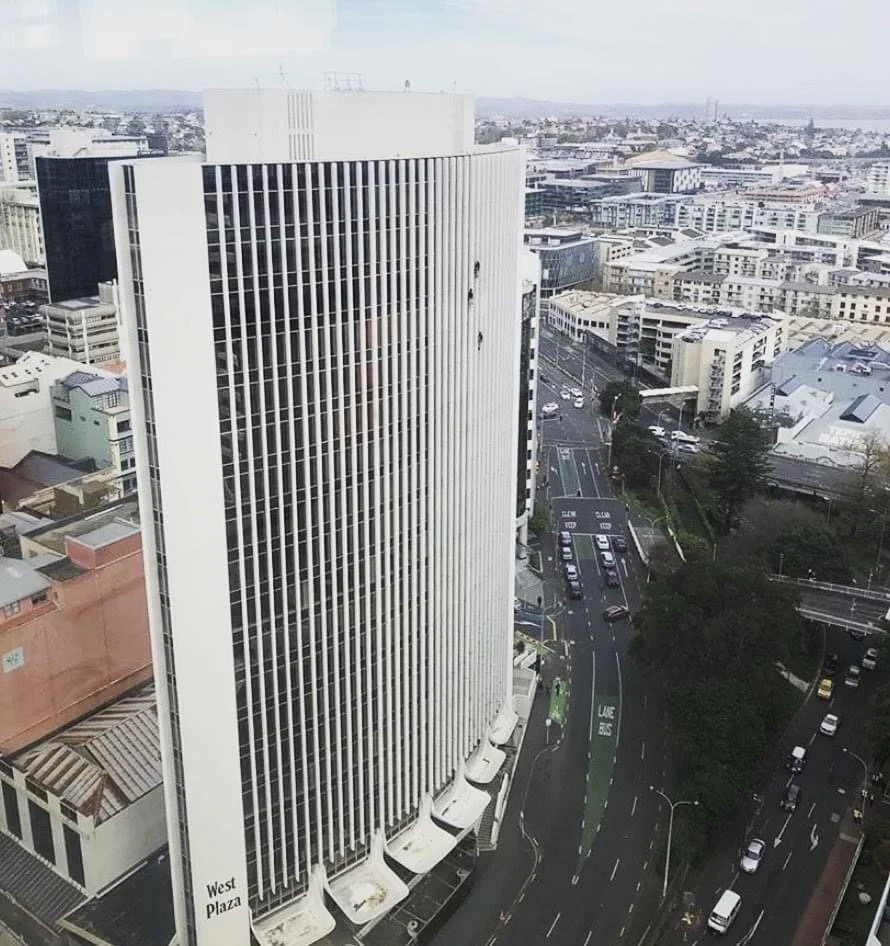 A tall white office building with vertical lines in an urban area, with surrounding roads, parked cars, and other buildings in the background.