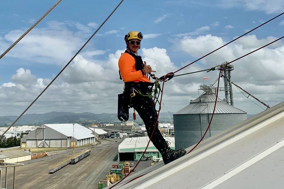 A worker in safety gear, including a helmet and harness, is perched on the edge of a roof, holding onto safety ropes with an industrial facility and mountains in the background.