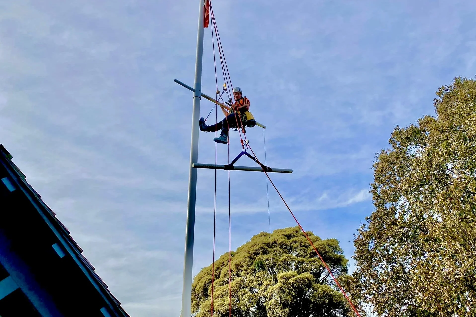 A person wearing a helmet and safety gear is sitting on a metal platform attached to a tall, vertical pole with ropes, appearing to be setting up or working on a TV antenna or similar structure against a partly cloudy sky, with trees visible in the b