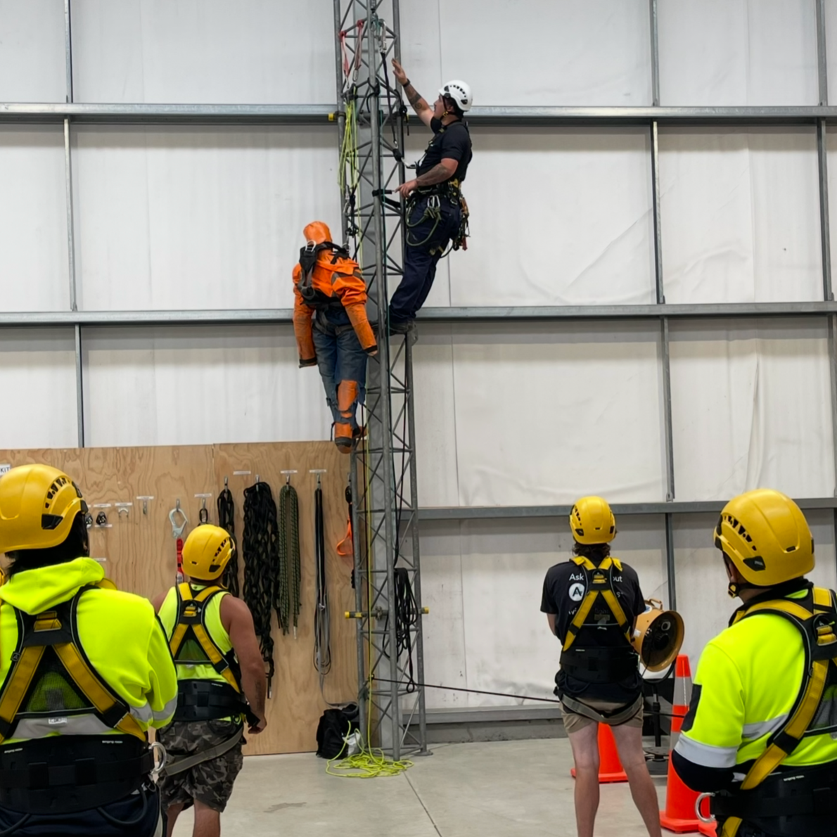 People in safety gear practicing climbing and rescue techniques on a metal tower inside a training facility.