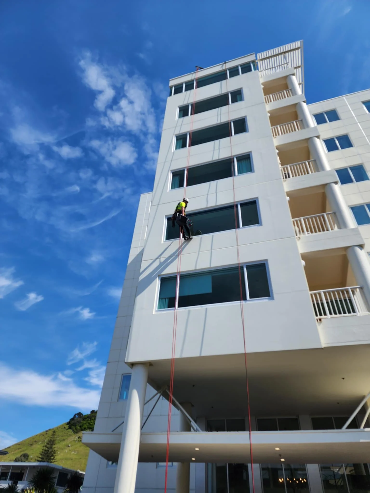 A worker in safety gear cleaning windows on a tall white residential building with balconies, under a partly cloudy blue sky.