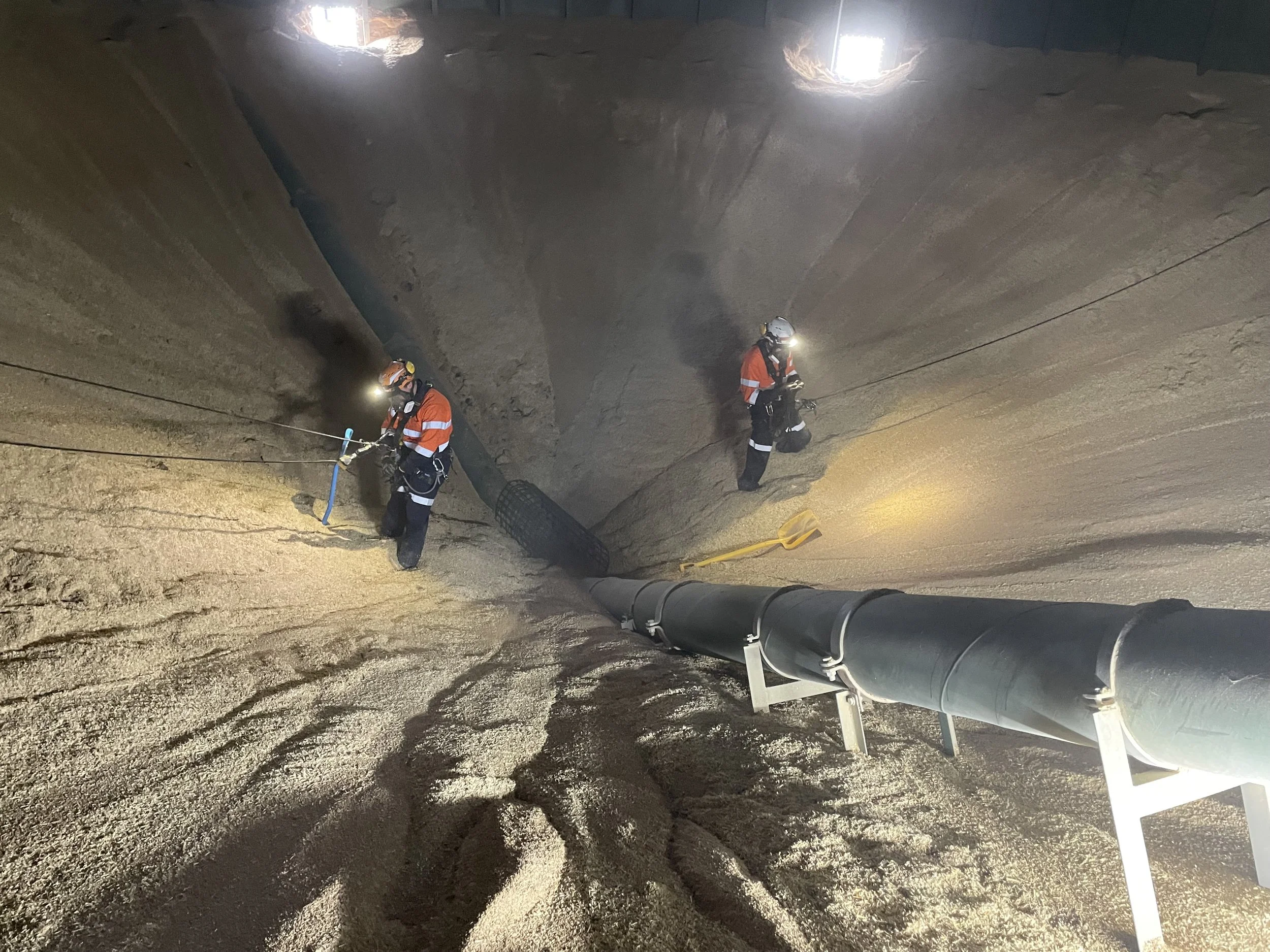 Two workers in orange safety vests and helmets inside a large industrial pipe or tunnel, inspecting or maintaining the pipe, with one holding a tool and both wearing headlamps for visibility.