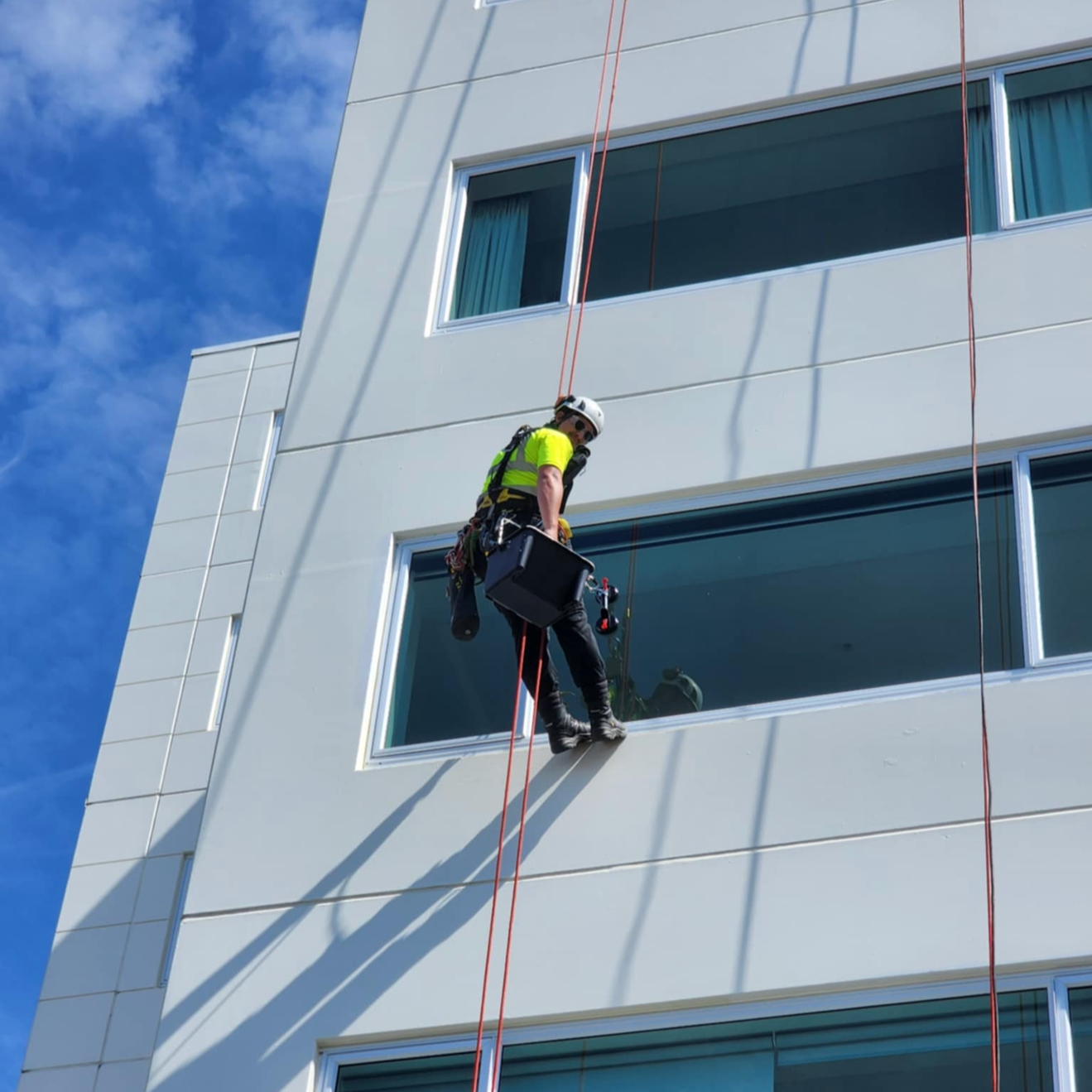 A rope access painter is suspended outside a modern white building, painting from a harness and ropes.