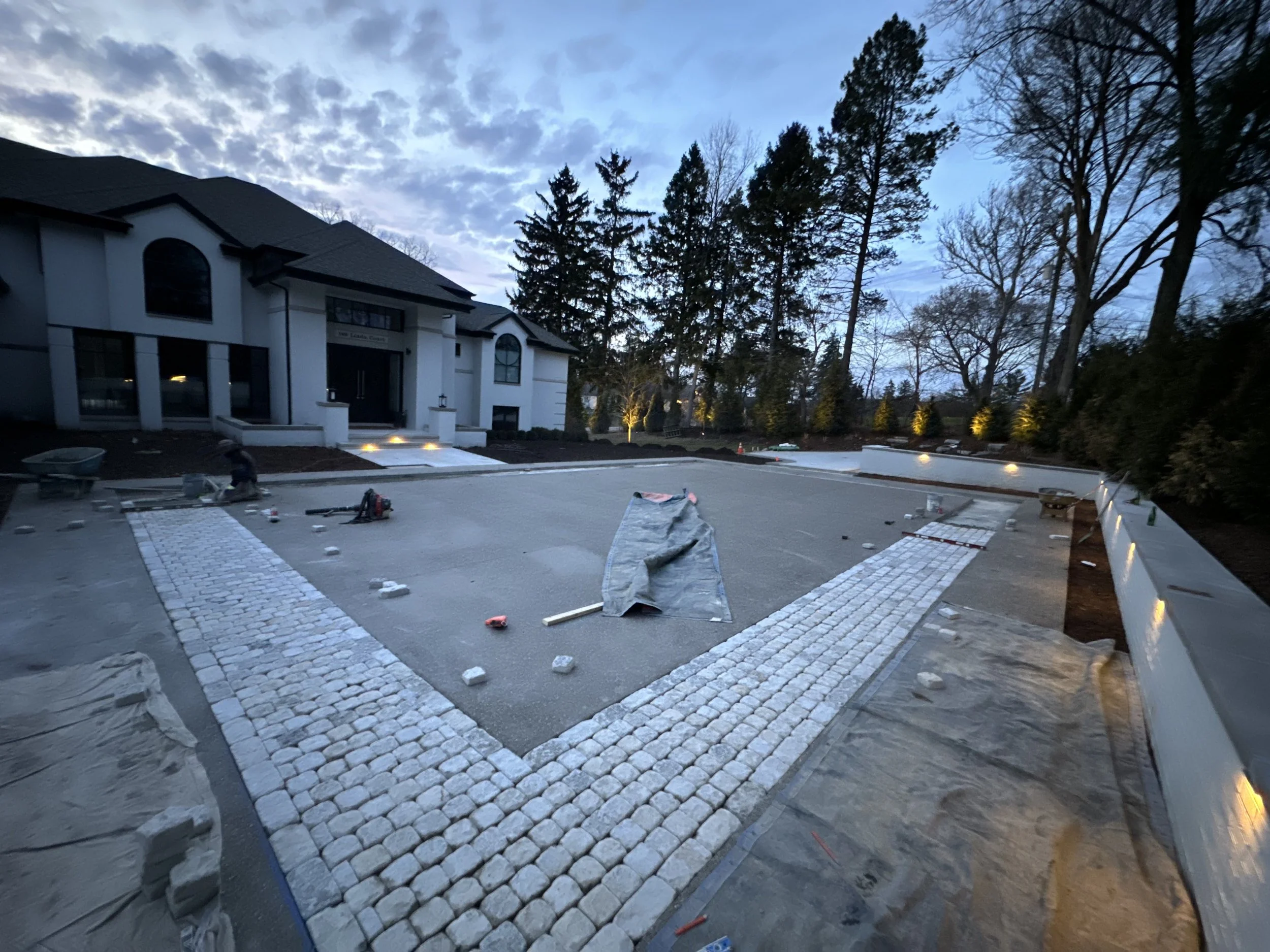 Driveway under construction with stone pavers, construction tools, and materials, in front of a large house with trees and shrubs on a clear evening.