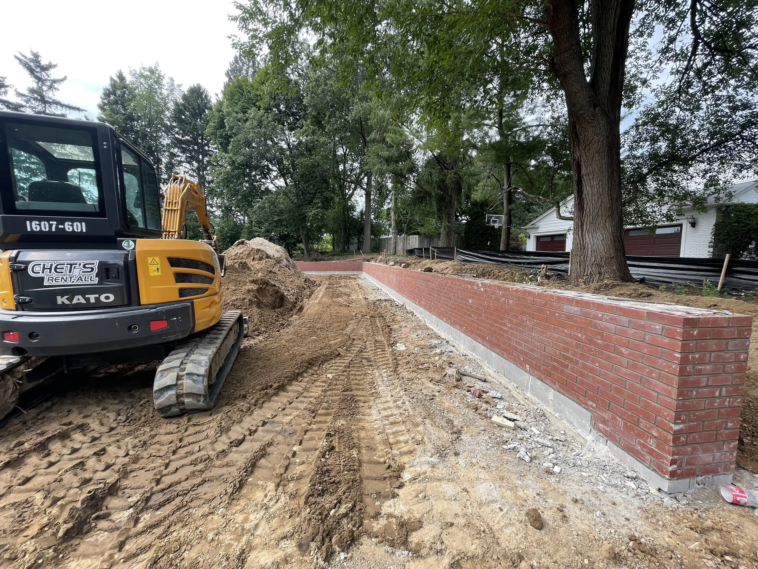 Construction site with a small excavator and a newly built brick wall, dirt and construction materials on the ground, trees and a house in the background.