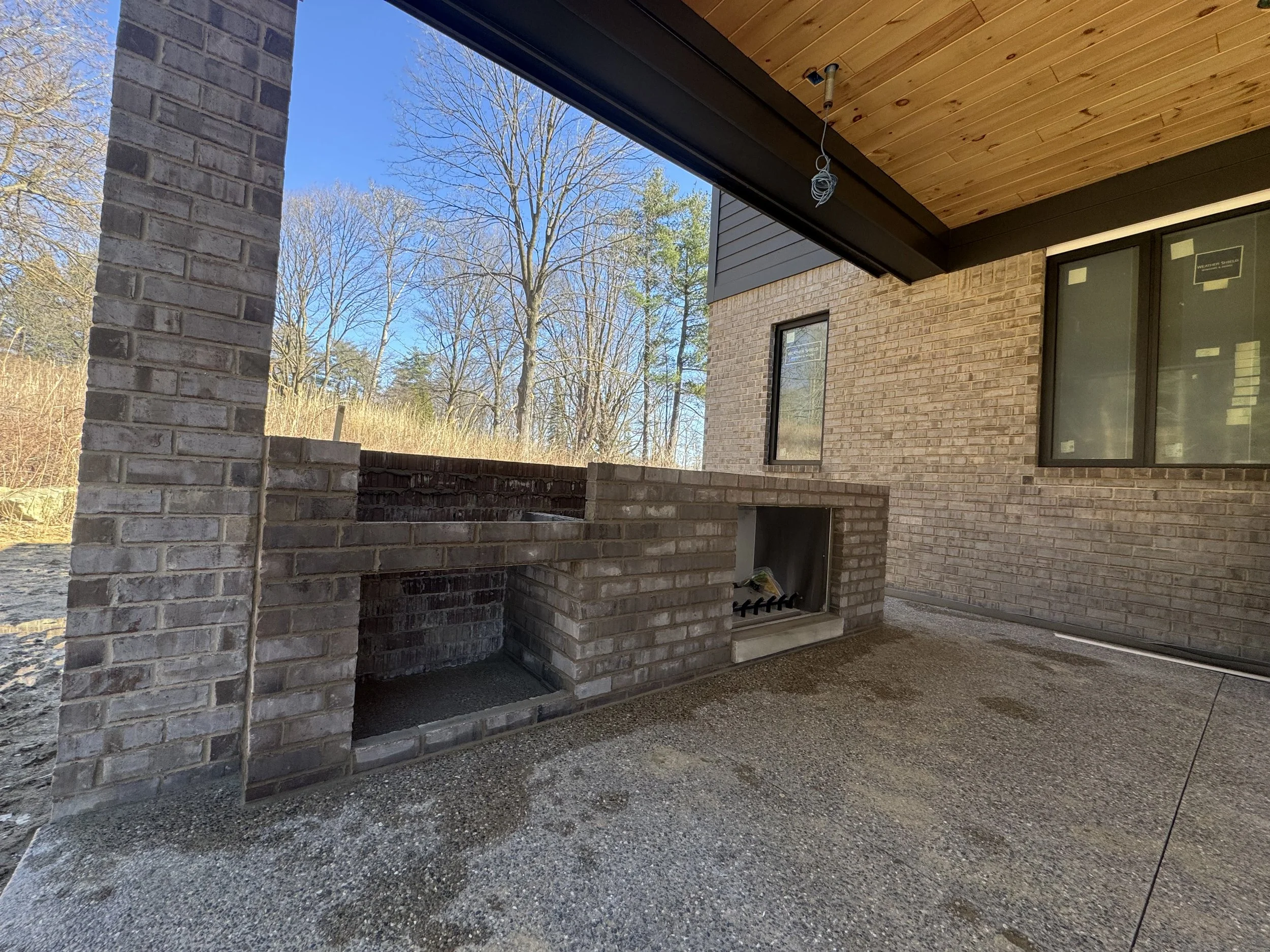 Part of a house under construction with brick walls, a concrete floor, and an outdoor brick fireplace or grill area, with leafless trees and blue sky in the background.