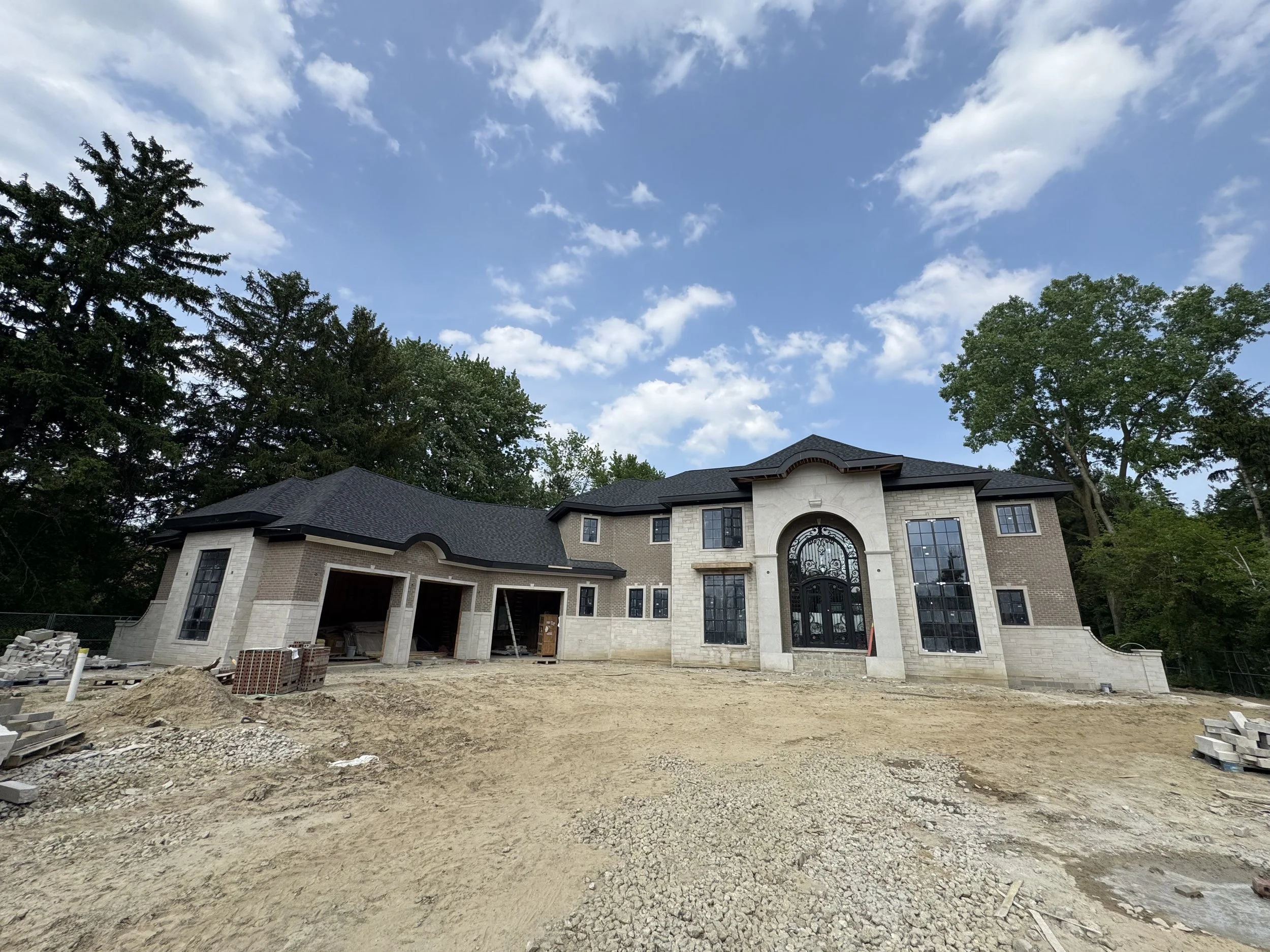 Under construction large house with a grand arched front door, tall windows, and a black roof, with a dirt yard and surrounded by trees and a partly cloudy sky.