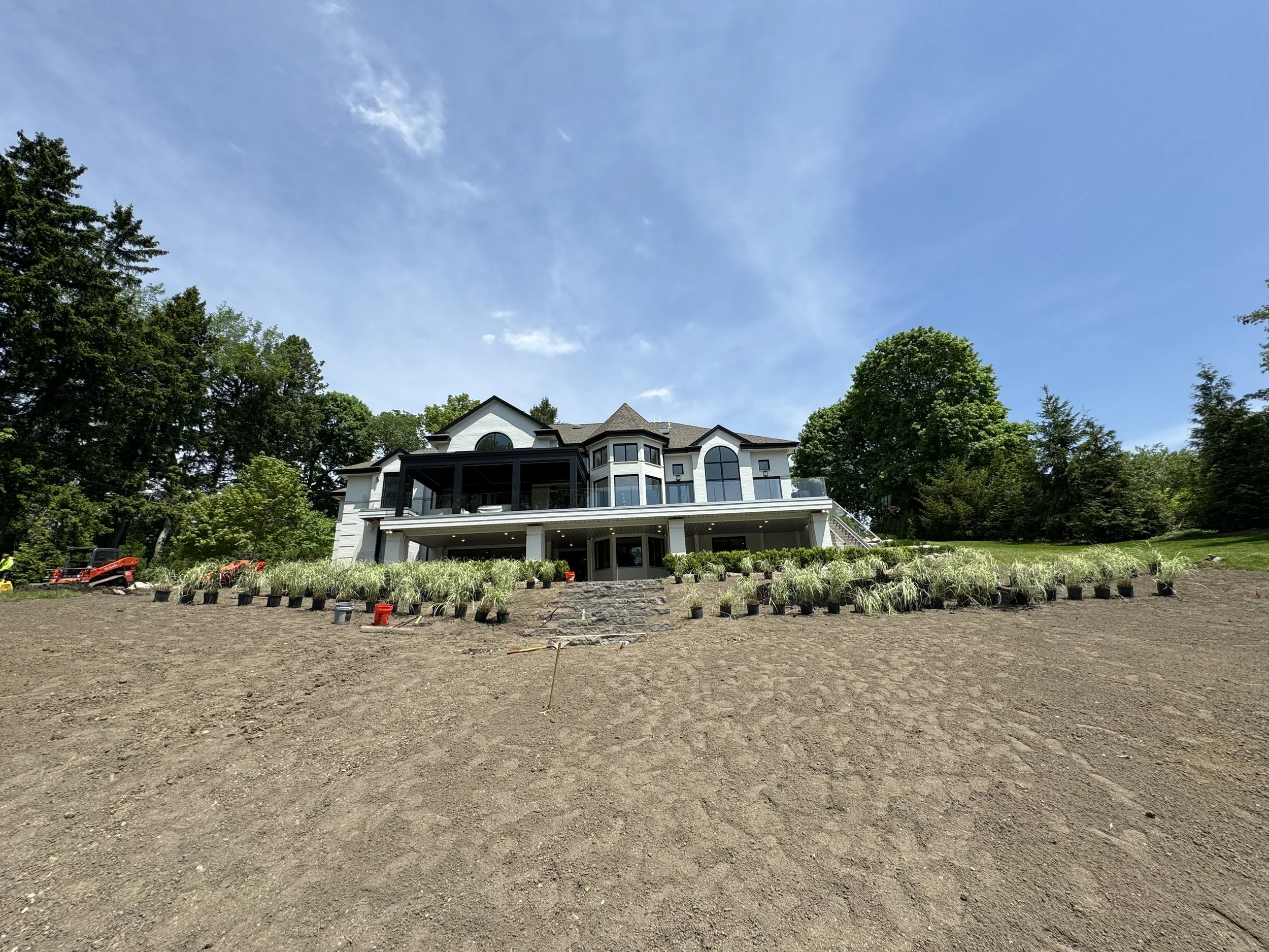 A large white house with multiple levels and arched windows, under construction, sits atop a hill with a cleared dirt yard in the foreground, surrounded by trees on a sunny day with a blue sky and a few clouds.