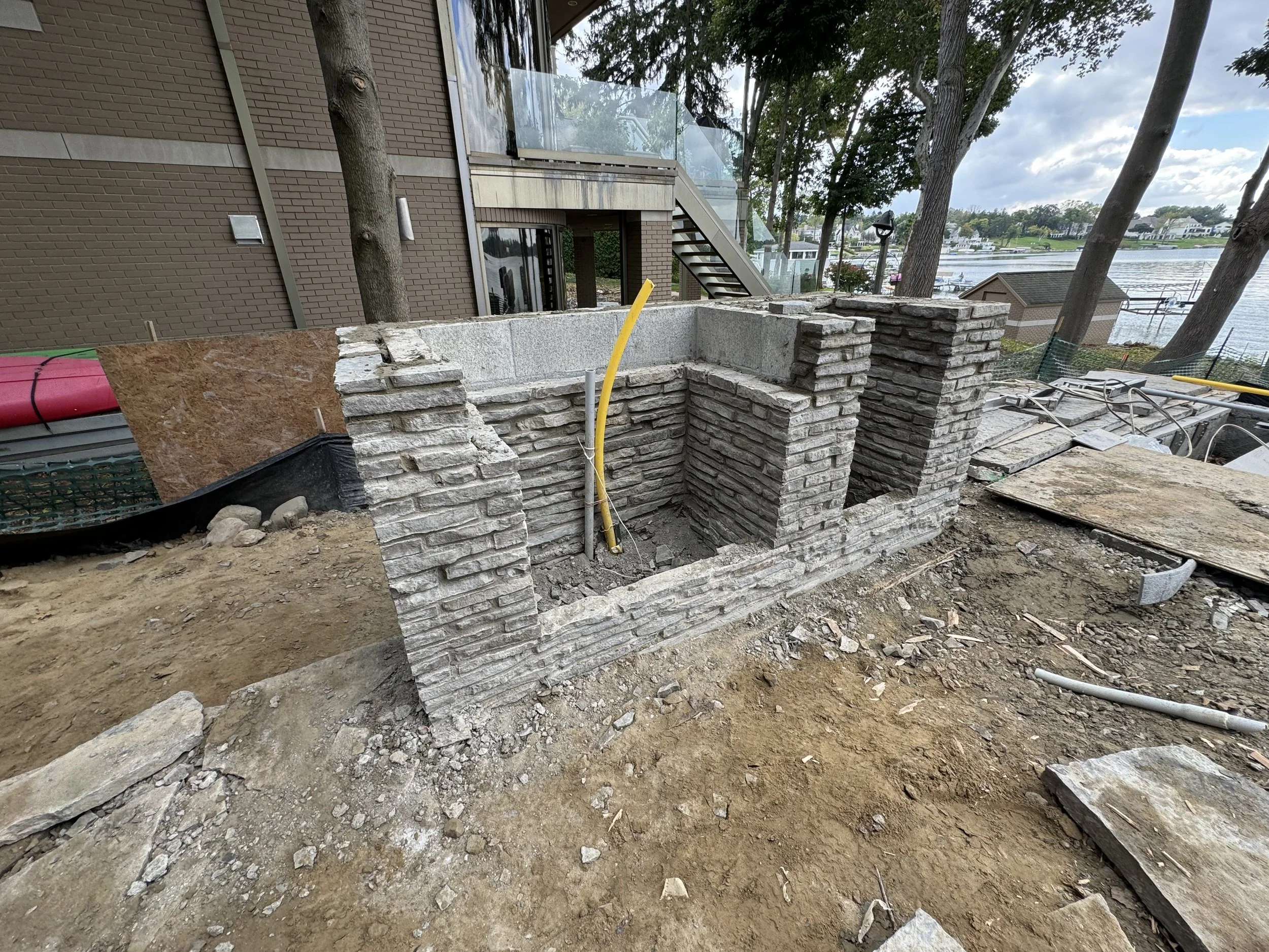 Under-construction outdoor stone BBQ structure, overlooking a body of water with trees and houses in the background.