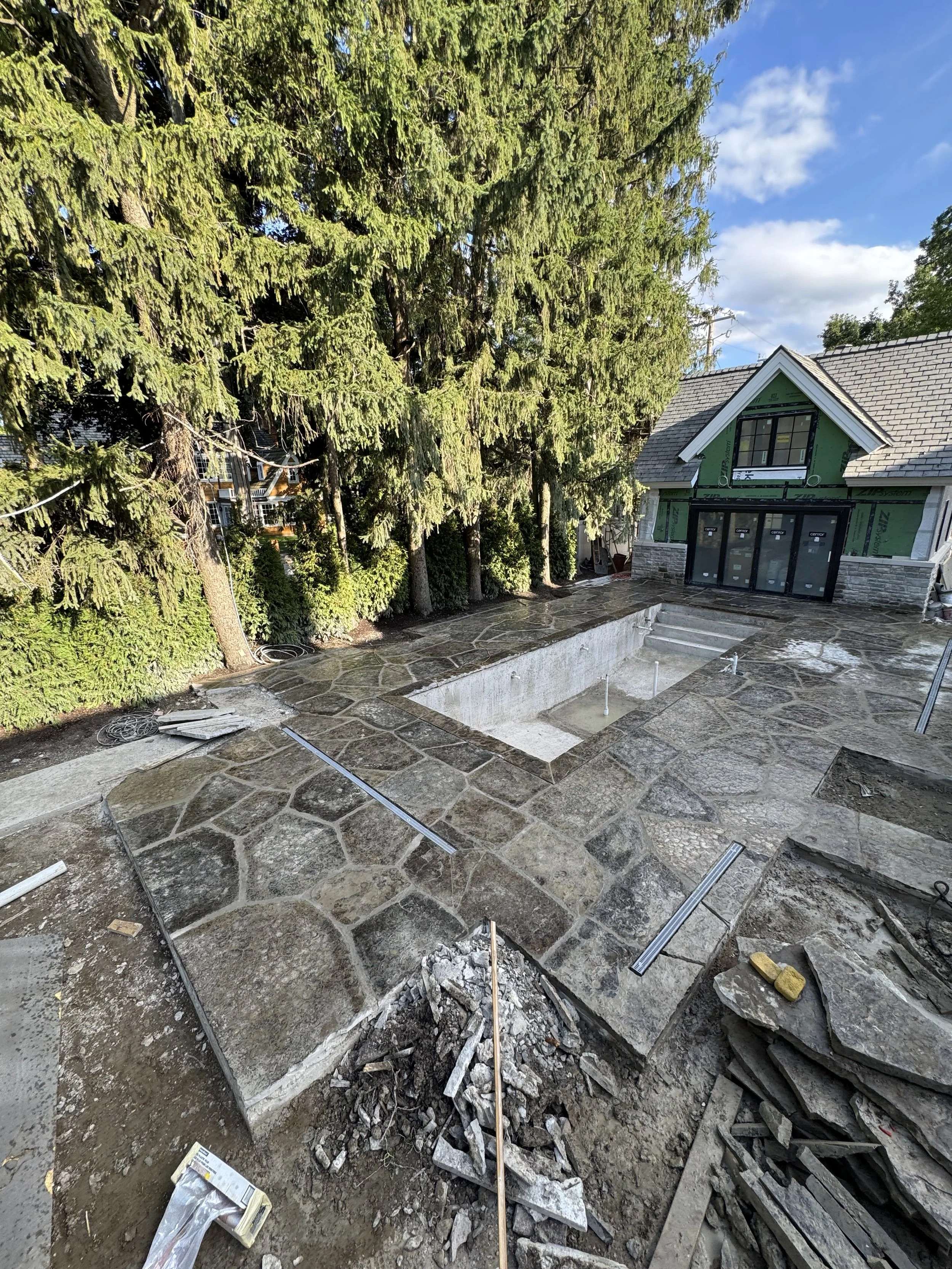 Construction site with stone patio, rectangular pool without water, and a house under renovation with scaffolding and green sheathing, surrounded by tall trees and cloudy sky.