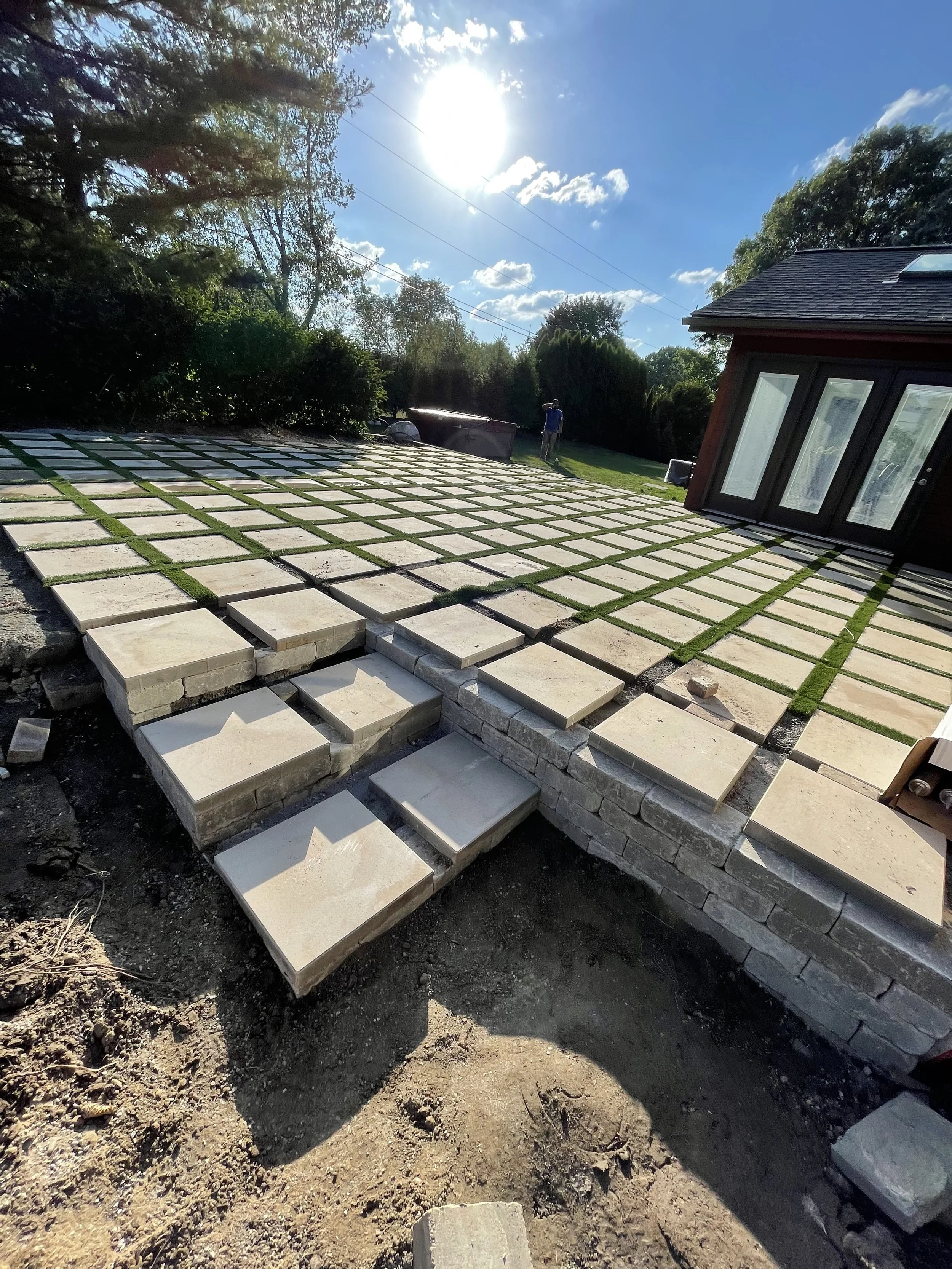 Paving stones being installed to create a new patio or pathway outside a house under a sunny sky.