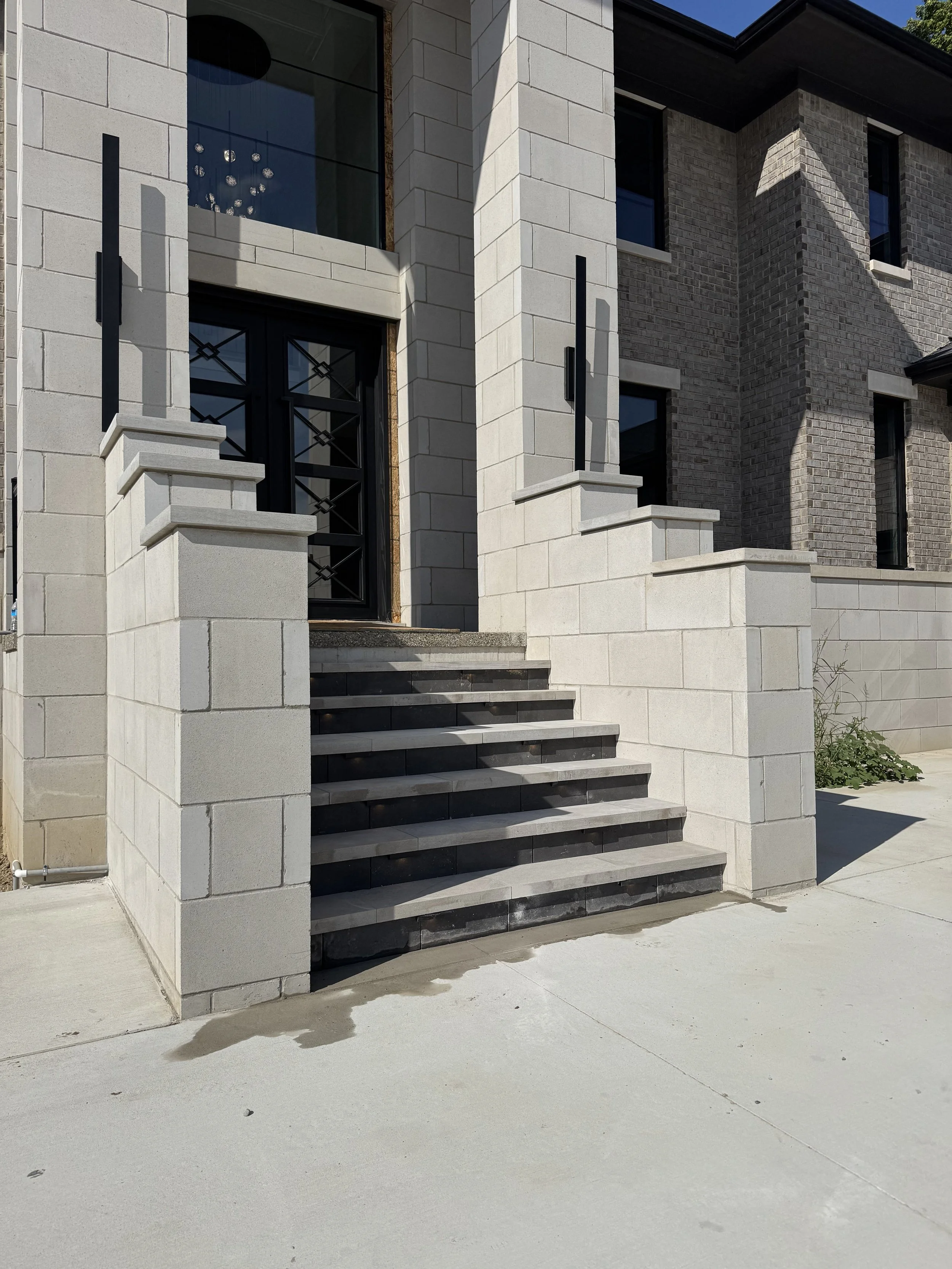 Exterior view of a modern residential building with stairs leading to the front door, featuring a mix of light and dark bricks, and decorative black railings.