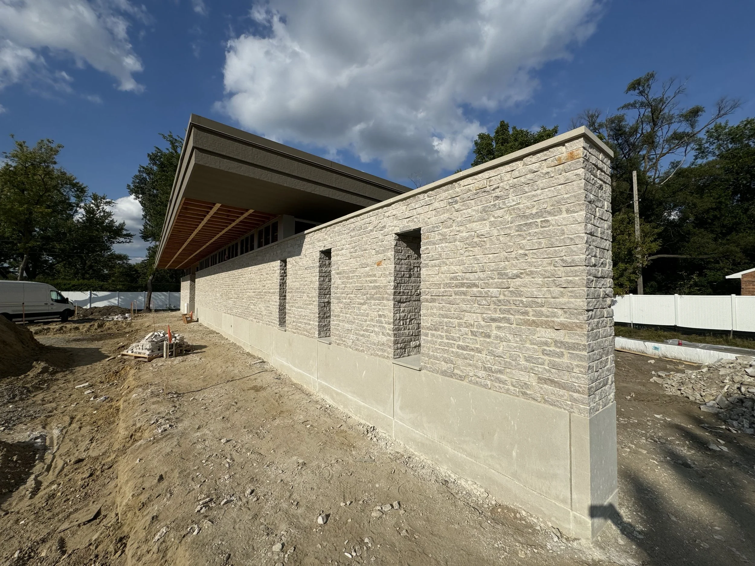 Construction site with a partially built cast-concrete and stone wall, dirt ground, construction materials, and a white fence in the background, under a partly cloudy sky.