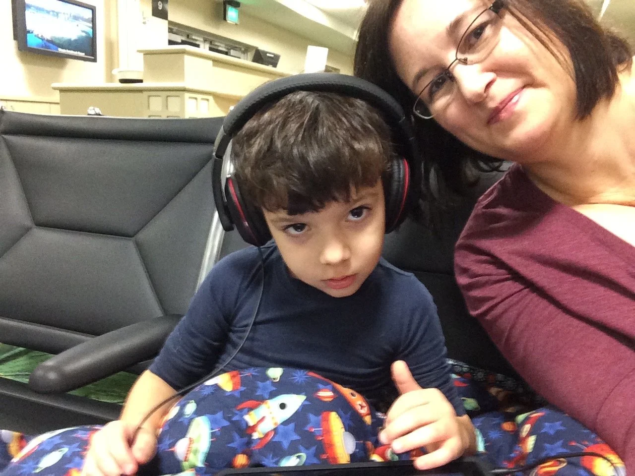 A woman and a young boy sitting in an airport waiting area taking a selfie. The boy wears large headphones and space-themed pajamas, and the woman is smiling.