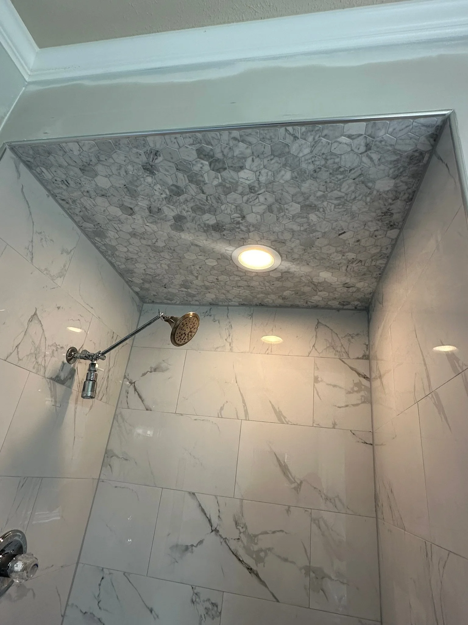 A bathroom shower area with large white marble tiles on the walls, a small round recessed ceiling light, and a shower head mounted on the wall. The ceiling has hexagonal marble tiles.