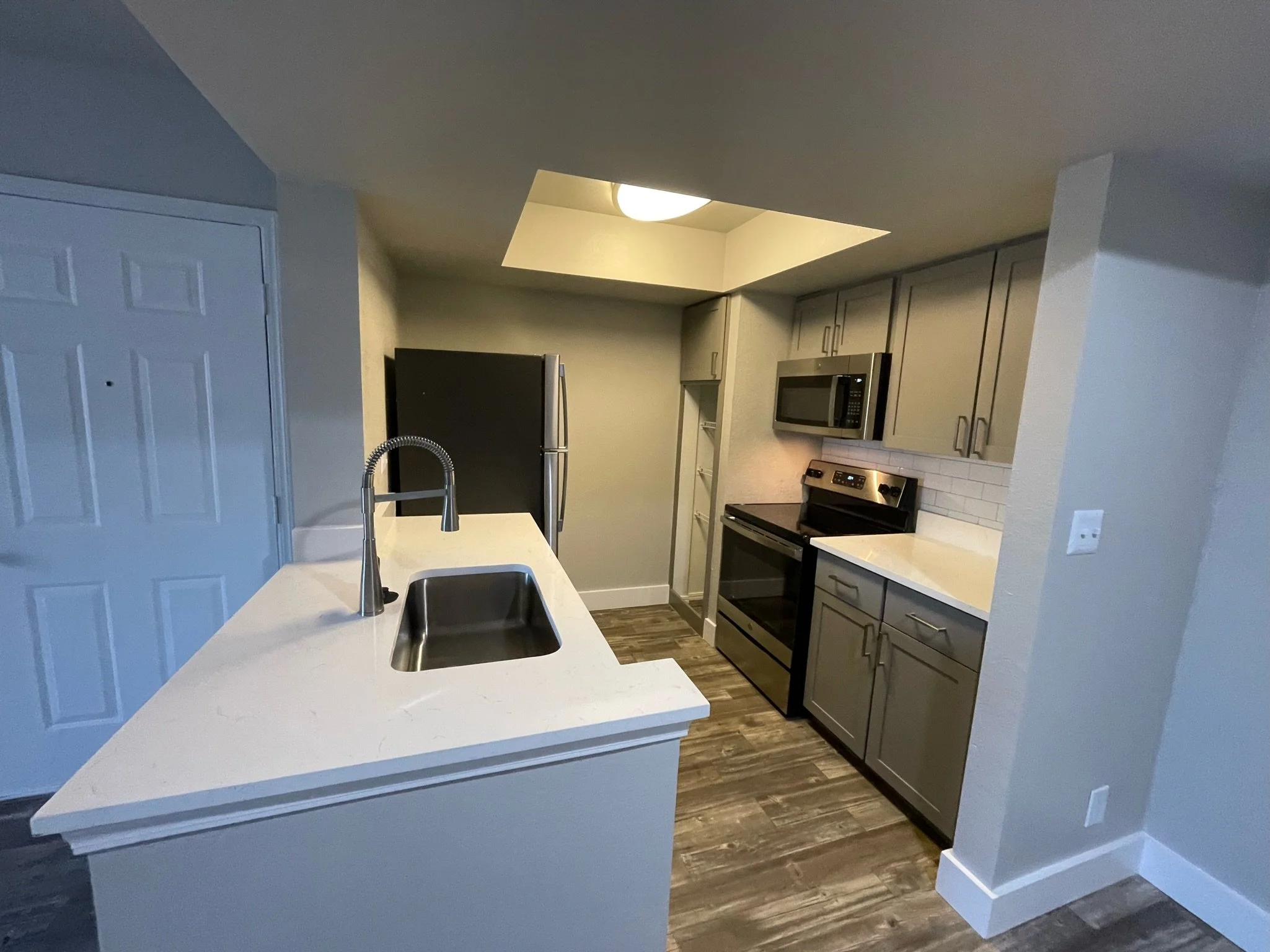 Modern small kitchen with gray cabinets, stainless steel appliances including a refrigerator, microwave, and stove, a white countertop with a sink, wooden flooring, and a small ceiling light.