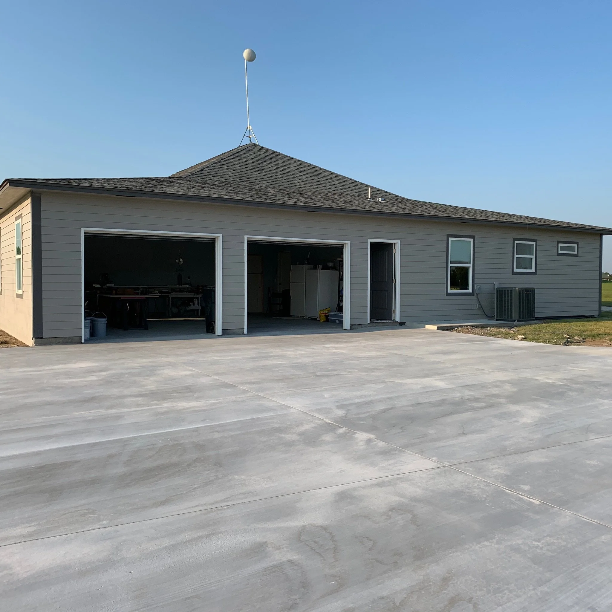 A house with a garage and a concrete driveway in front. The garage doors are open, revealing inside appliances and furniture. The house has gray siding and a dark roof, with a blue sky background.