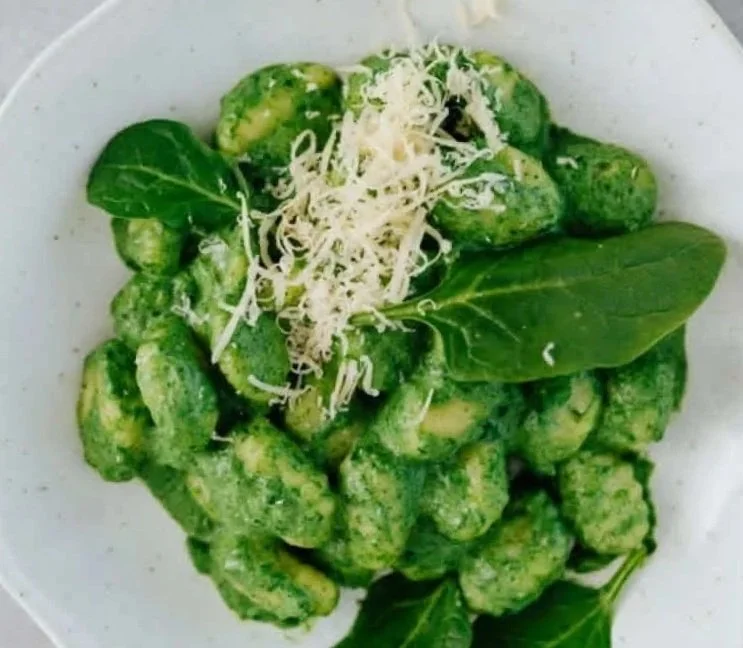 A bowl of green gnocchi with shredded cheese and basil leaves on top.