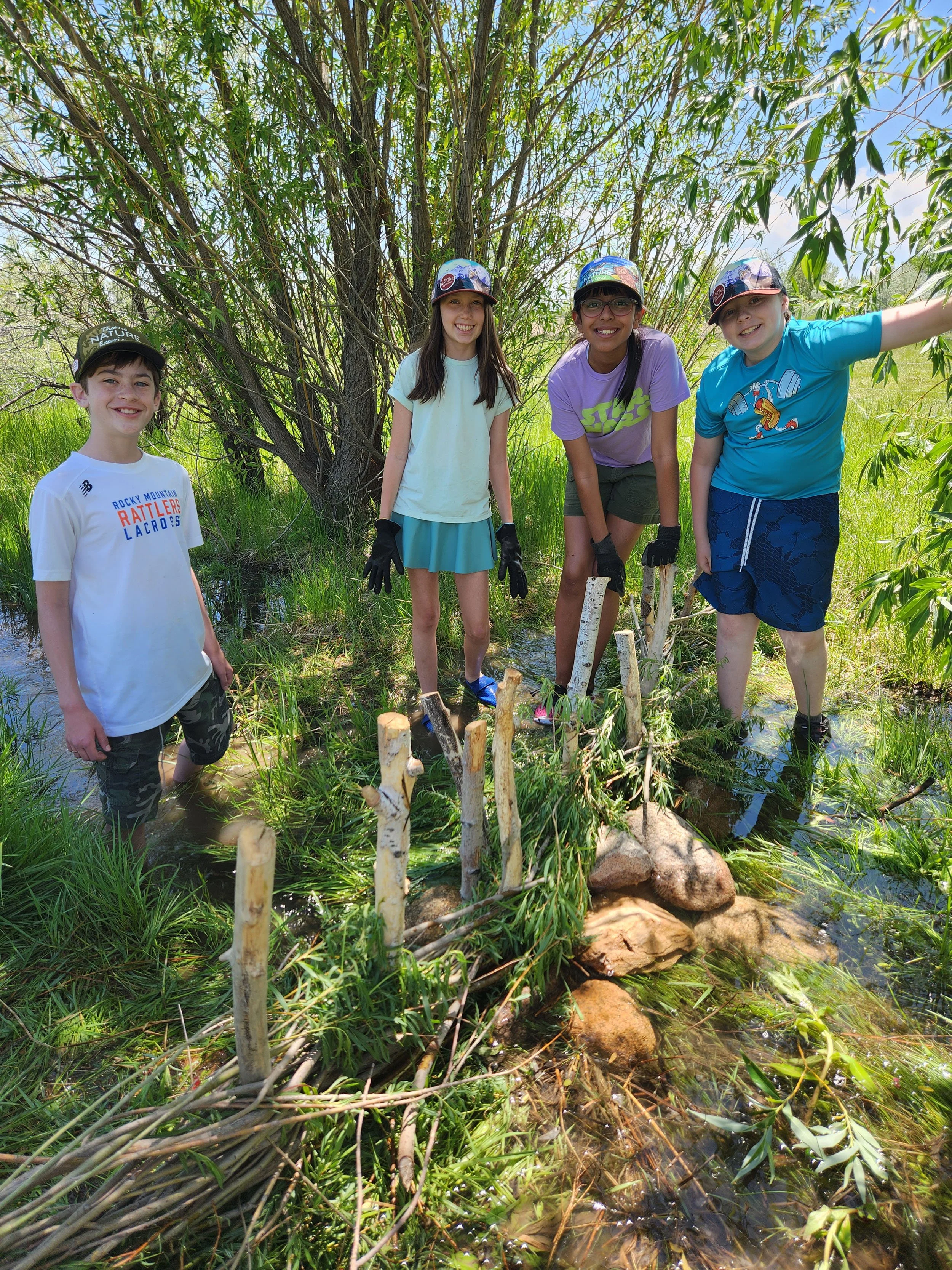 Four children standing in a marshy area with tall grass, trees, and rocks, holding sticks and wearing gloves, enjoying an outdoor nature exploration.