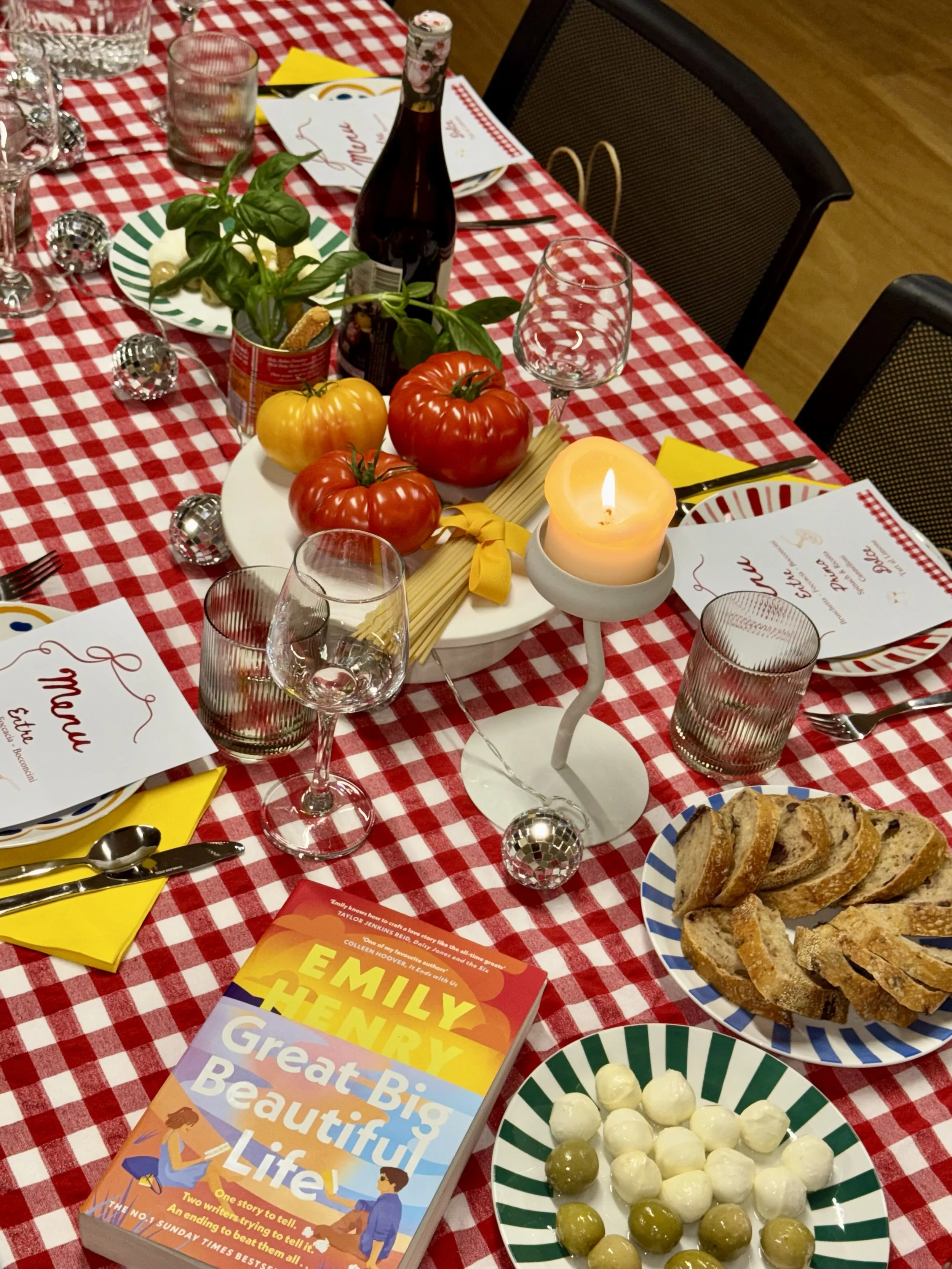 A festive table setting with a red and white checkered tablecloth features a white plate with fresh tomatoes, a plate with sliced bread, a striped bowl of cheese balls, a striped bowl of olives, a candle, a wine bottle, glasses, a potted herb, and a book titled 'Great Big Beautiful Life' on the table.