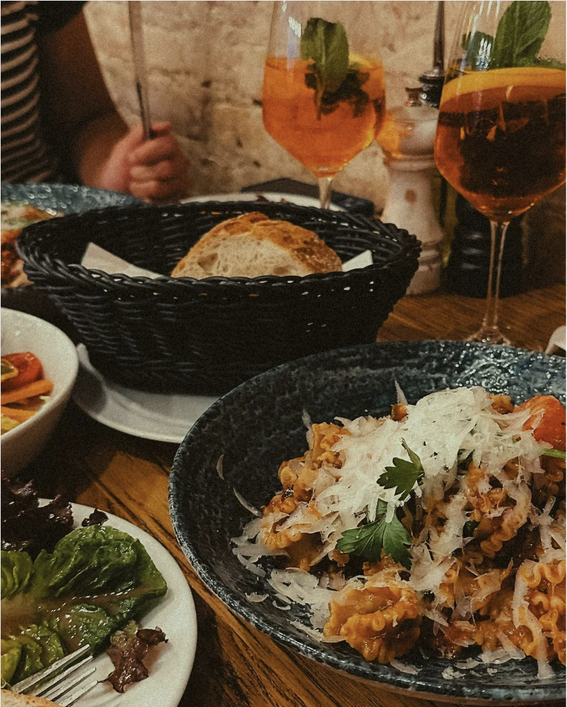 A table with a bowl of pasta garnished with herbs, a basket of bread, two glasses of orange-colored drinks with mint and lemon garnish, and a small dish of green vegetables.