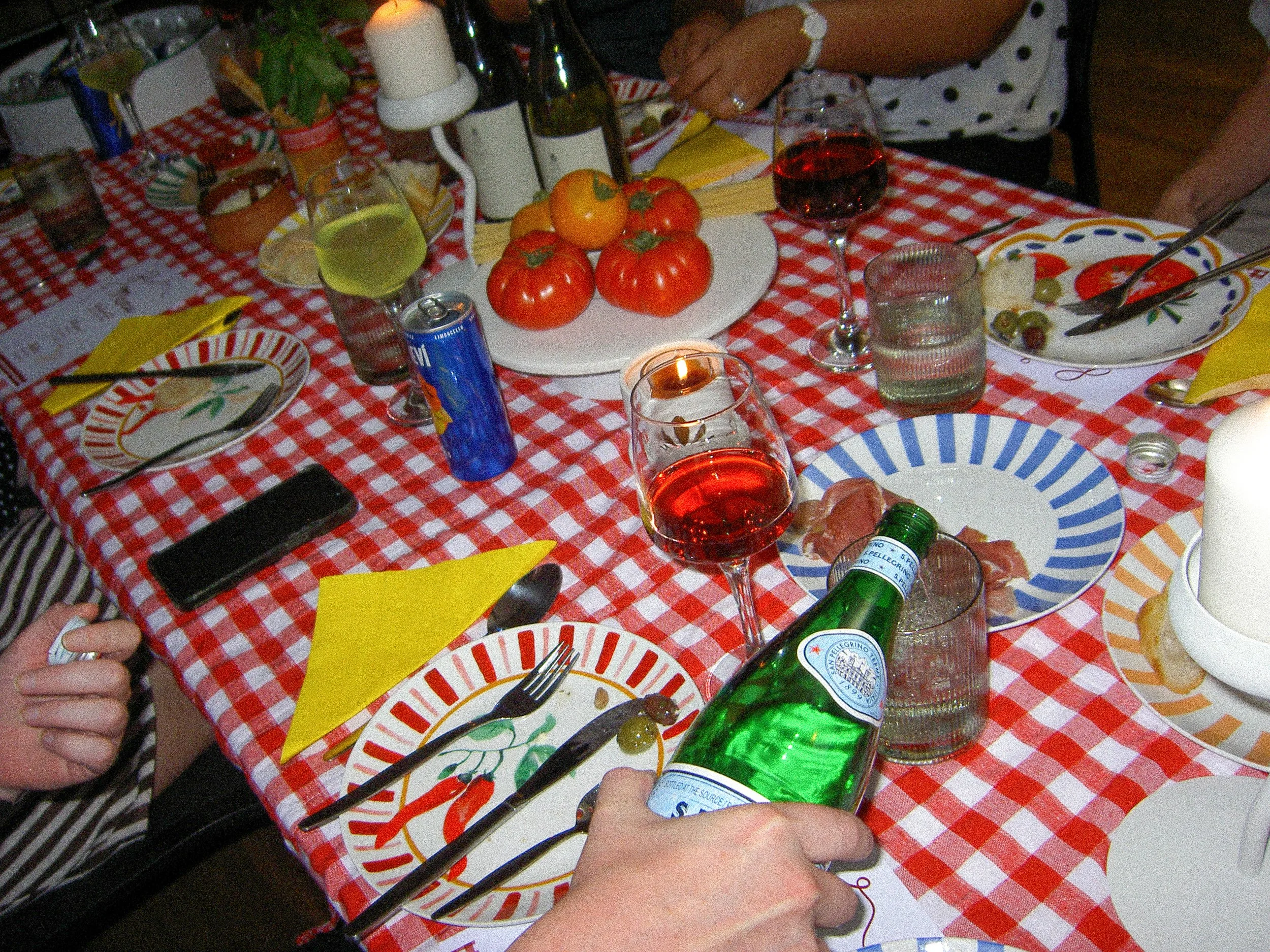 A dinner table with a red and white checkered tablecloth, set with plates, glasses of wine, bottles, a candle, and a centerpiece of tomatoes and other vegetables.