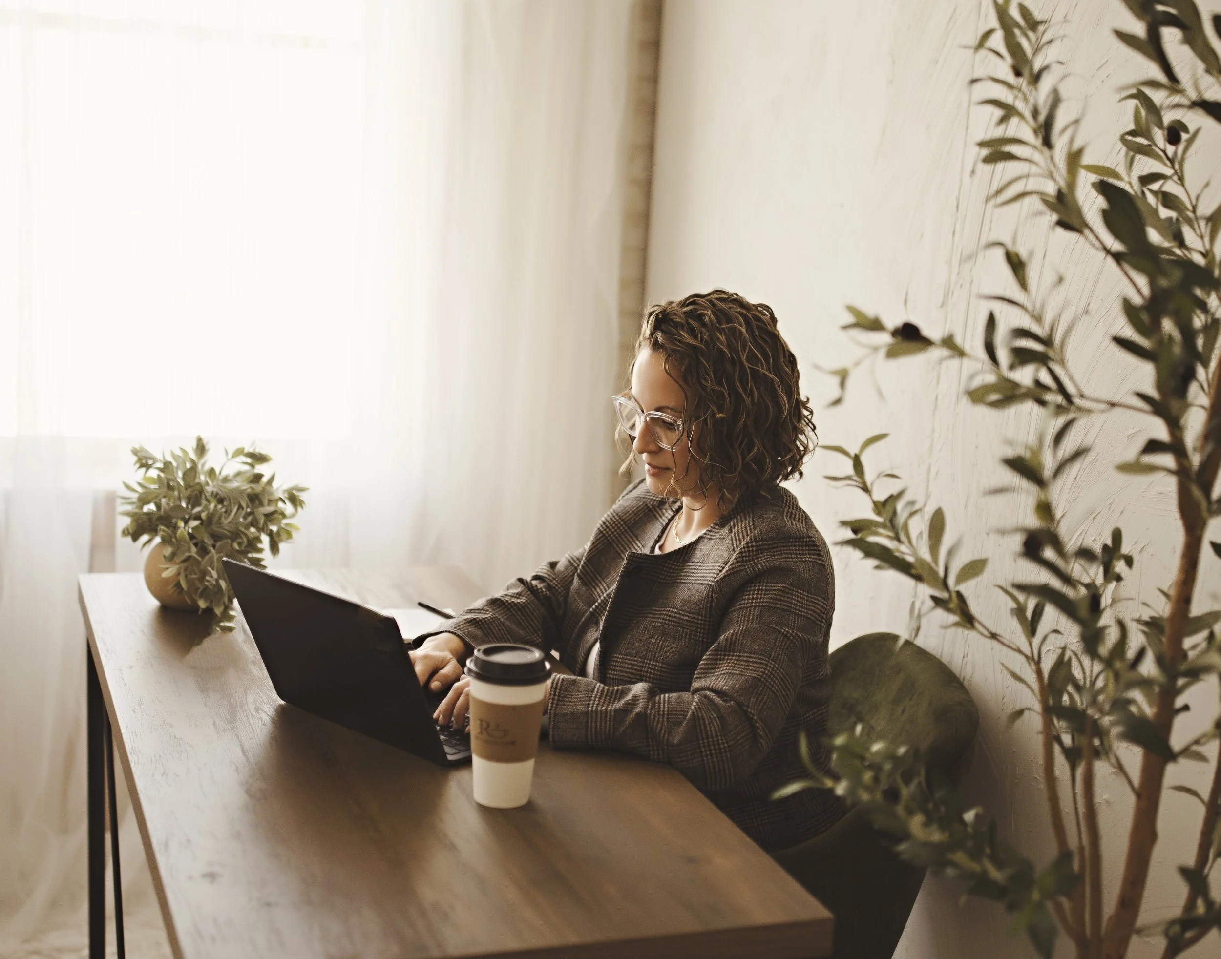 A woman with curly hair and glasses working on a laptop at a wooden desk with a plant and a coffee cup in a well-lit room.