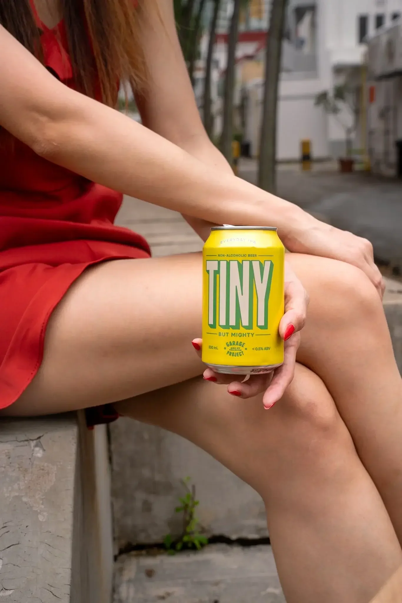 A person in red shorts sitting outdoors holding a yellow can of non-alcoholic beer.