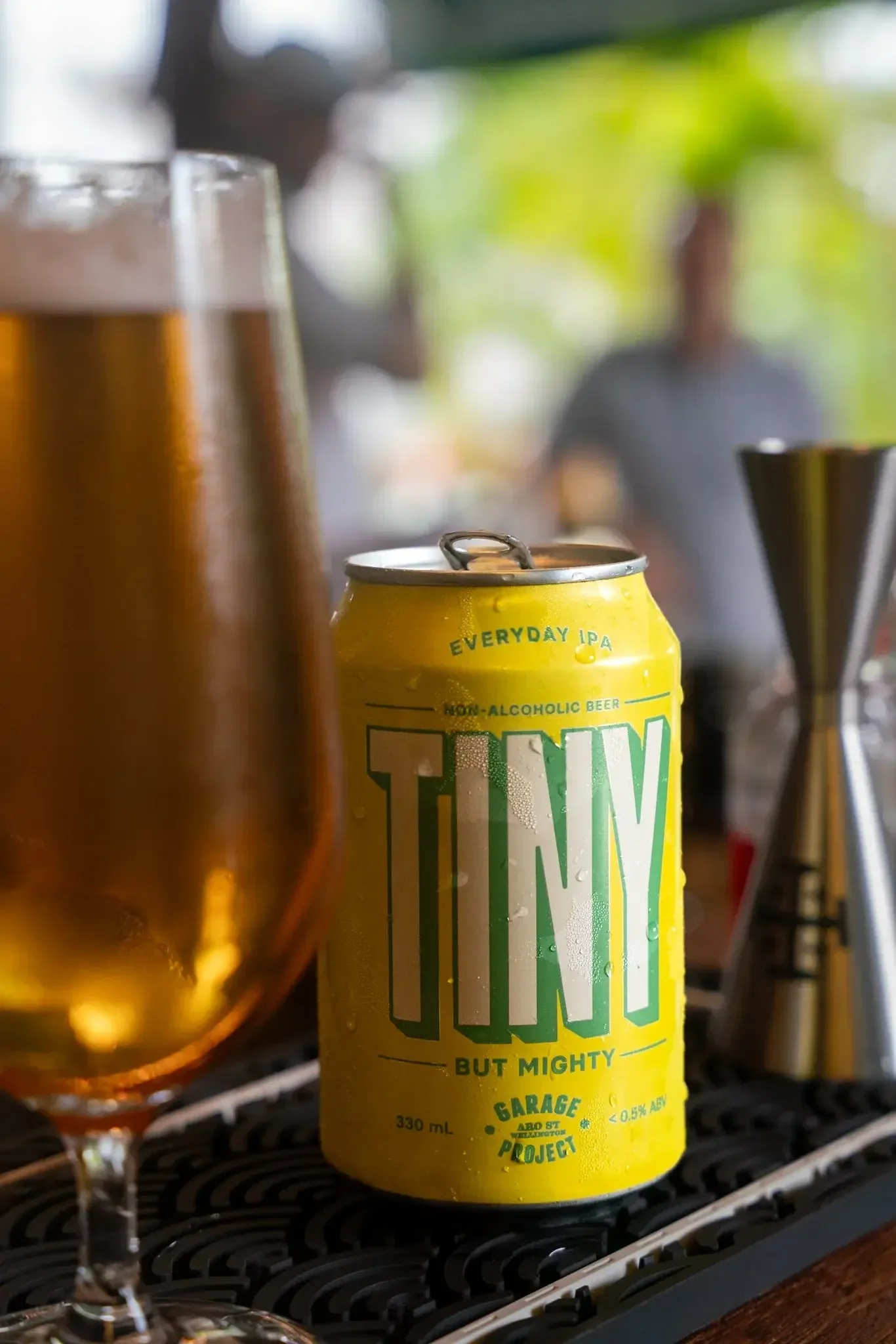 A yellow can of non-alcoholic beer with green text, placed on a black grill surface, with a blurred background of a person and green foliage.
