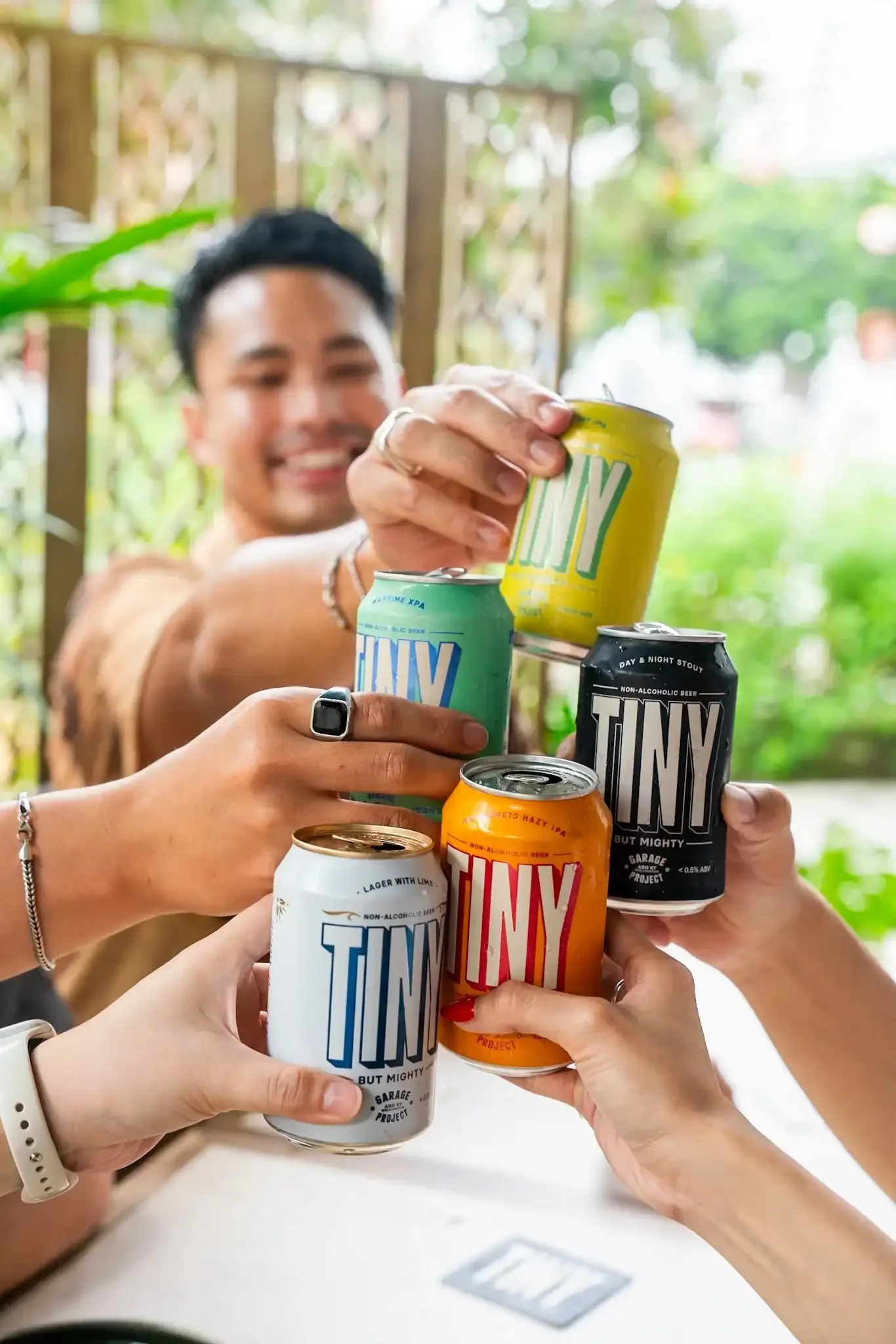 Multiple hands holding cans of TINY non-alcoholic beer in various flavors, with a smiling man and a blurred outdoor background.