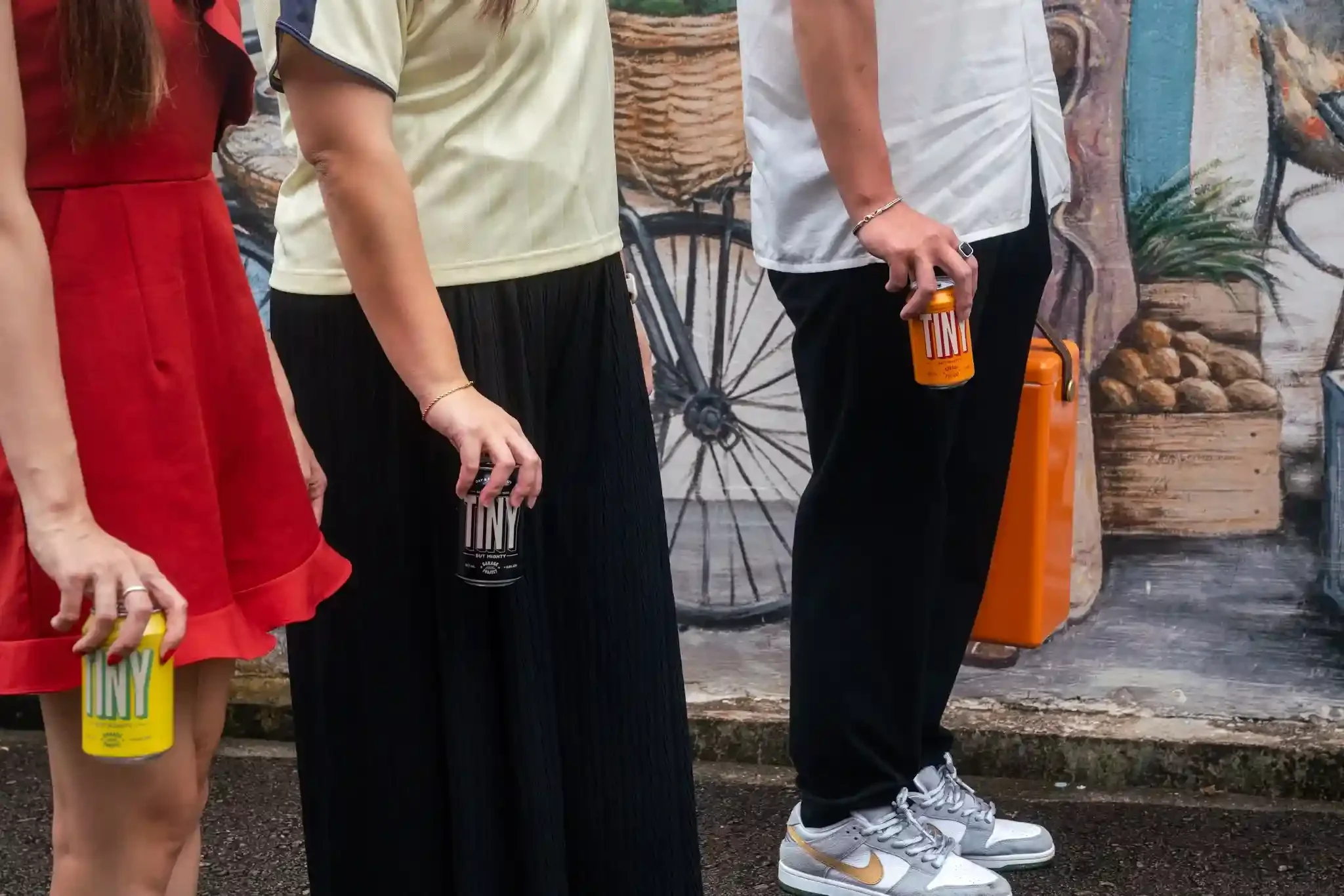Close-up of the lower halves of four people standing in a line, each holding a can of TINY Beer, with a city mural background featuring a bicycle and plants.