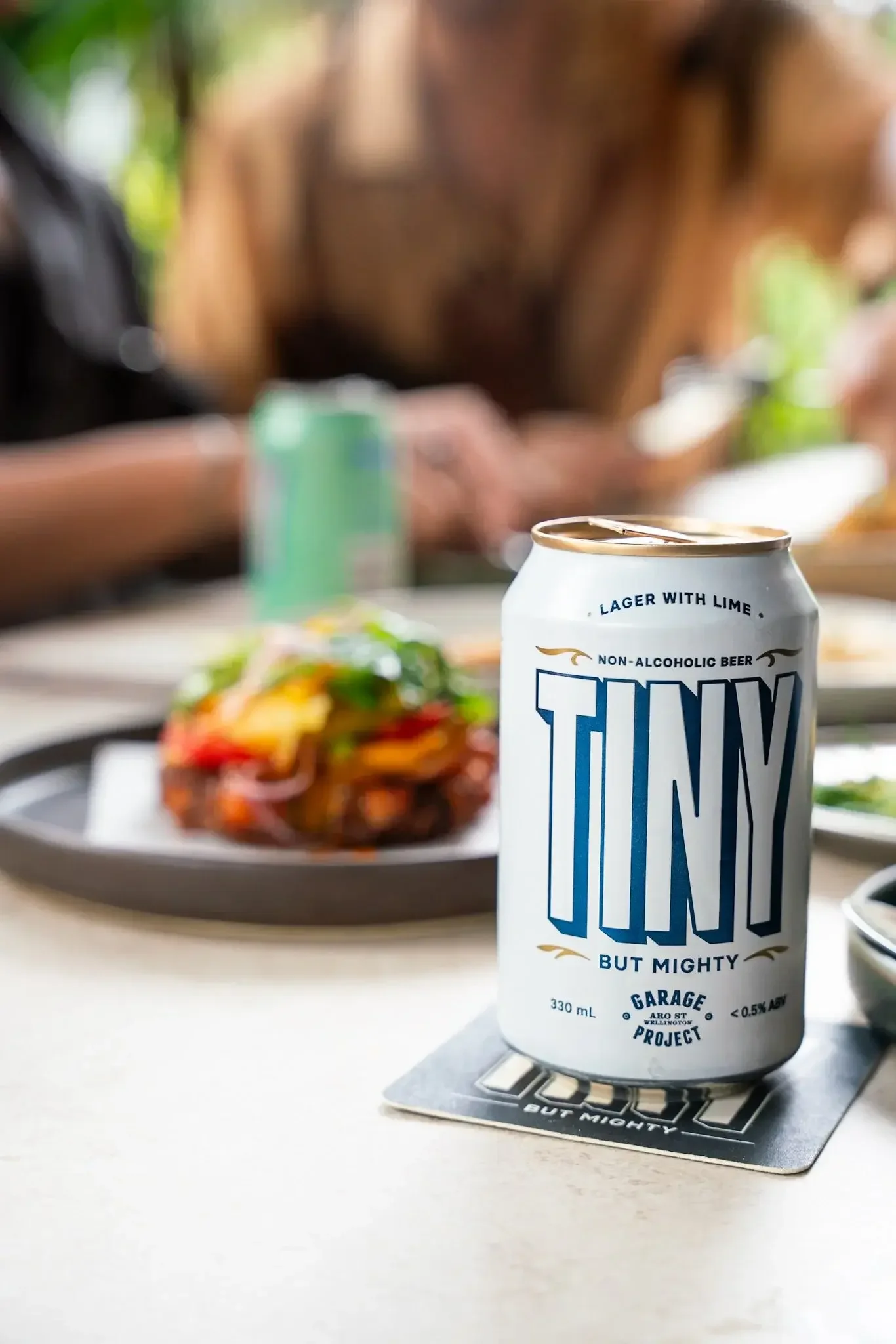 A white can of non-alcoholic beer with blue and gold text and design, placed on a coaster on a table. In the background, there is a blurred plate of food and a person with long hair sitting at the table.